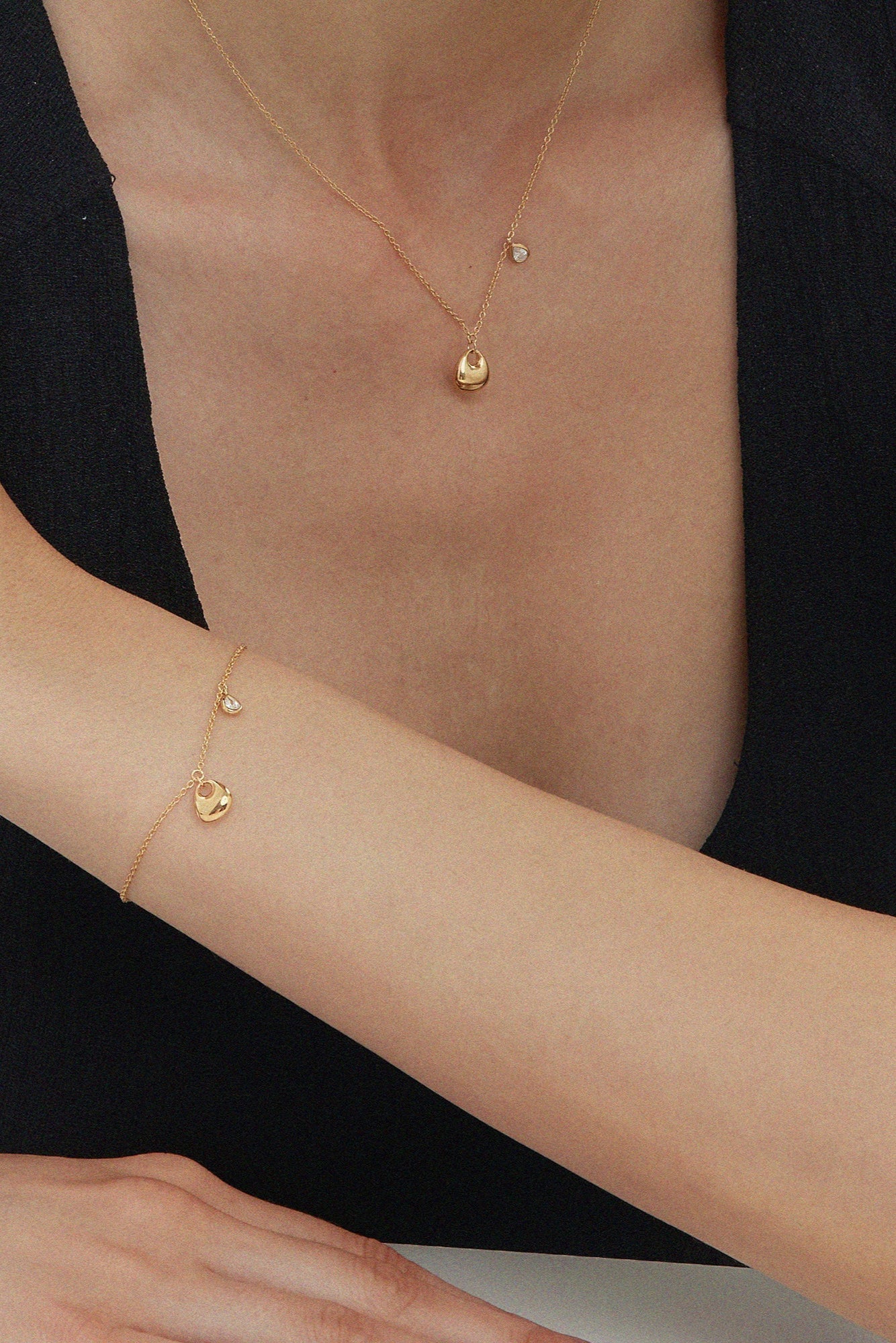 A woman in a sleeveless black top rests her arm on a surface, wearing delicate gold jewelry, including the Nora Double Pendant Necklace in Gold, plus a matching bracelet and ring. Her other hand gently touches her shoulder against a white background.