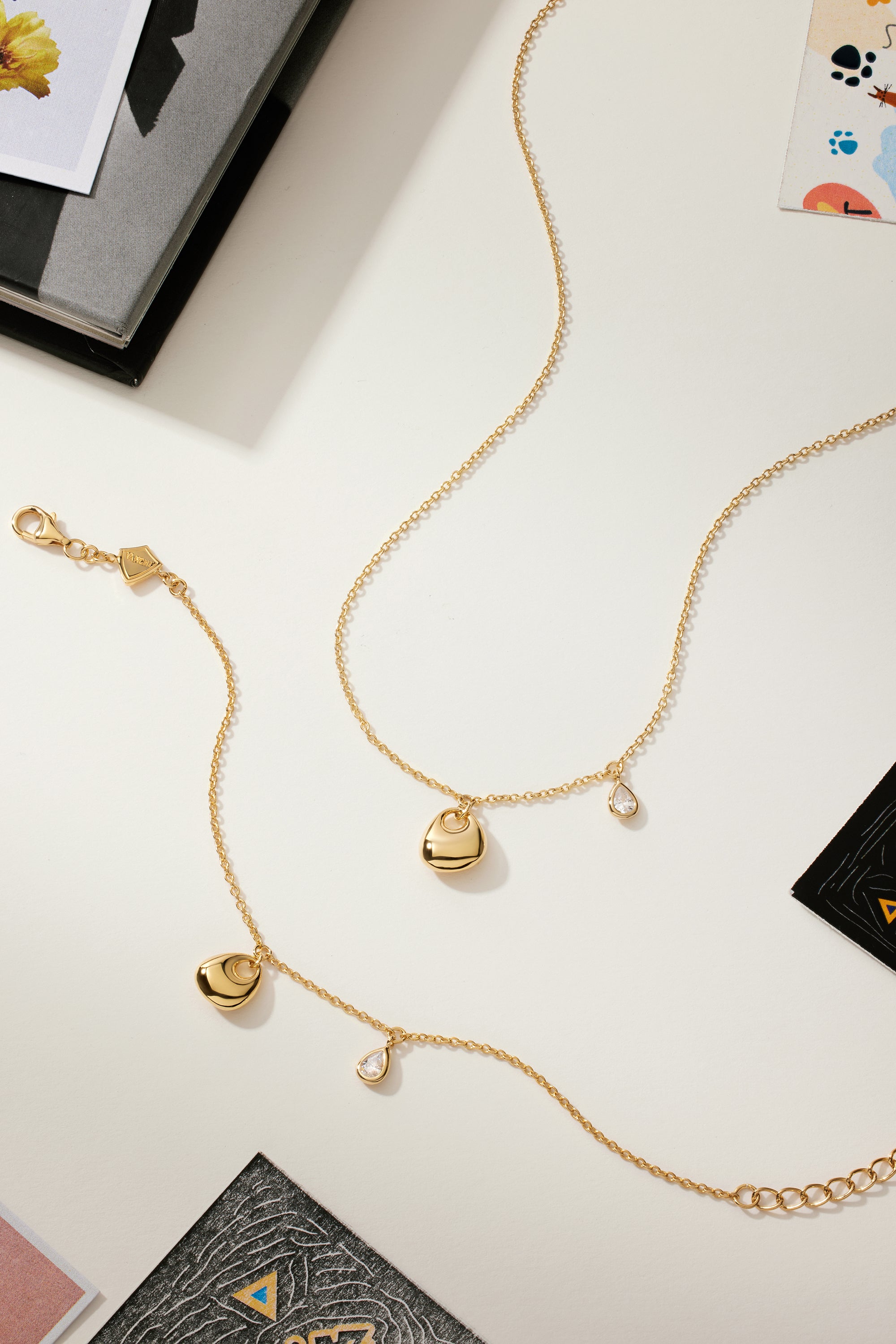 The Nora Double Pendant Necklace in Gold, featuring small sculptural charms such as a teardrop-shaped cubic zirconia, is displayed on a white surface surrounded by scattered colorful papers and cards.
