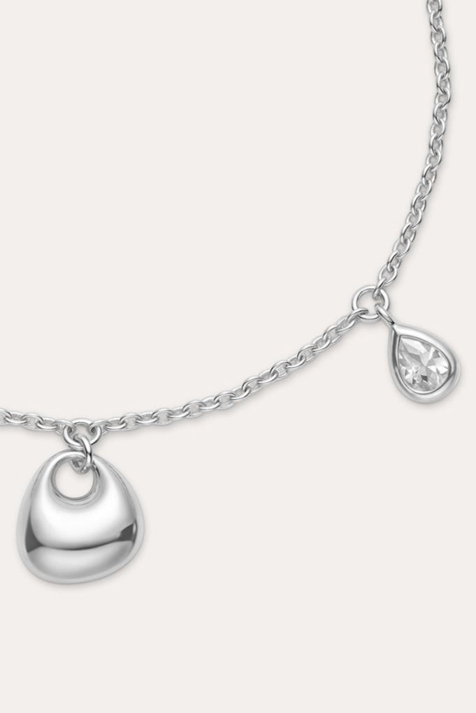 The Nora Double Pendant Bracelet in Silver features an adjustable clasp, a small heart-shaped charm, and a tiny cubic zirconia teardrop charm dangling side by side.