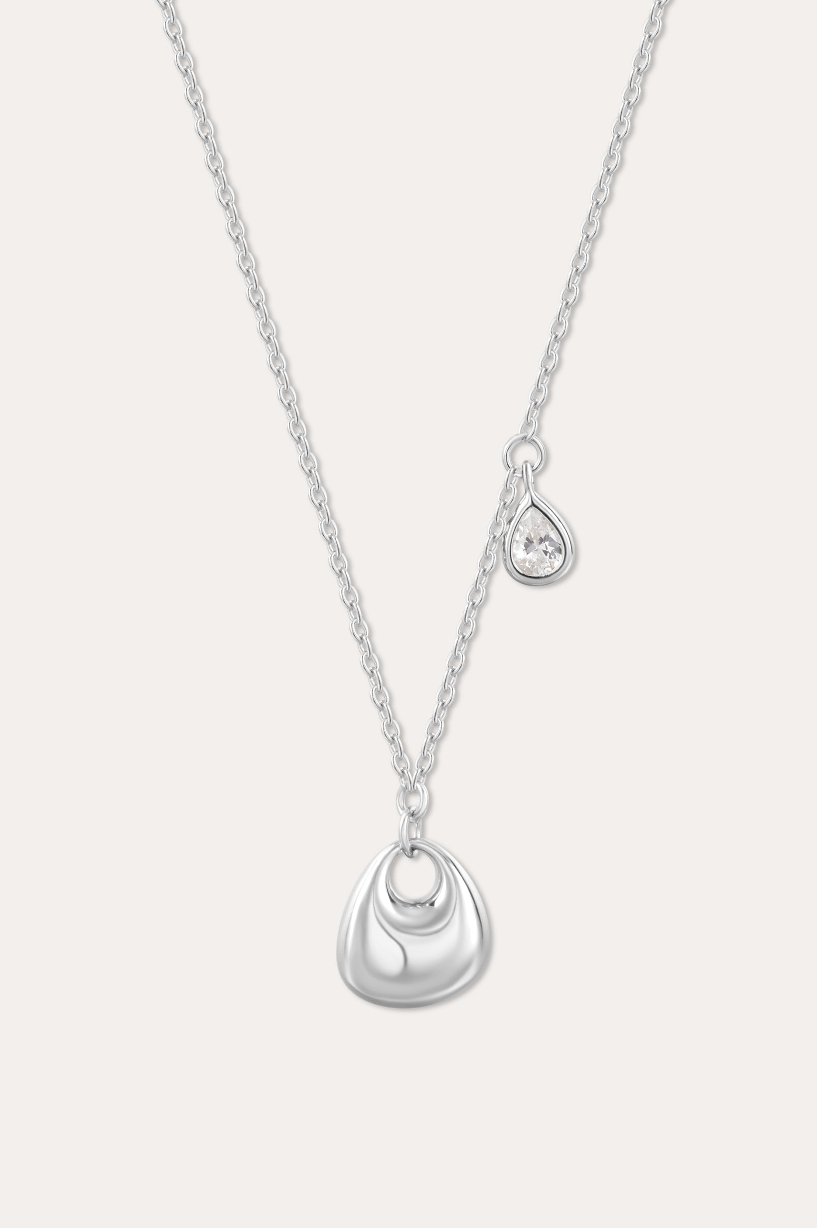 The Nora Double Pendant Necklace in Silver features a fine chain with a shiny sculptural pendant and a separate tiny teardrop charm set with a clear cubic zirconia, displayed against a plain background.