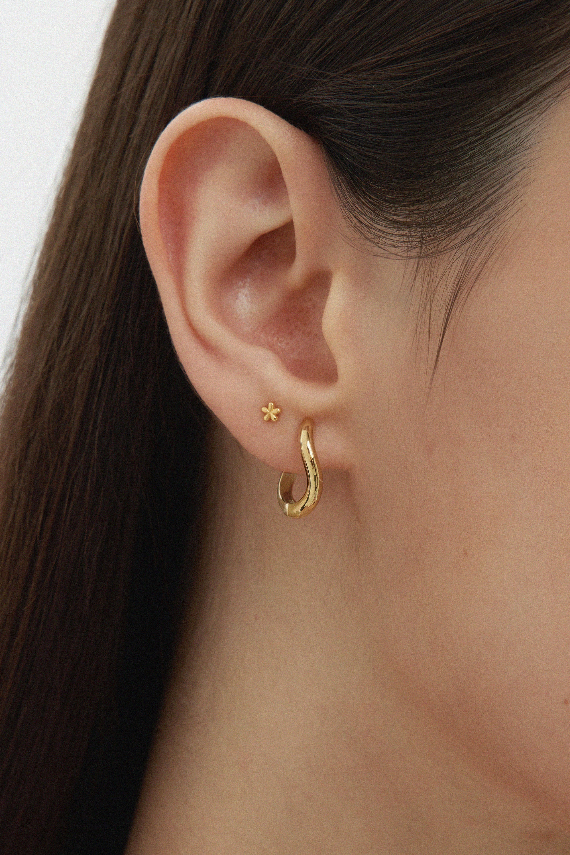 Close-up of a womans ear adorned with two earrings: a small flower stud and the Nori Twisted Hoop Earrings in Gold. The neutral background highlights the elegant shine of these 18k gold plated hoops.