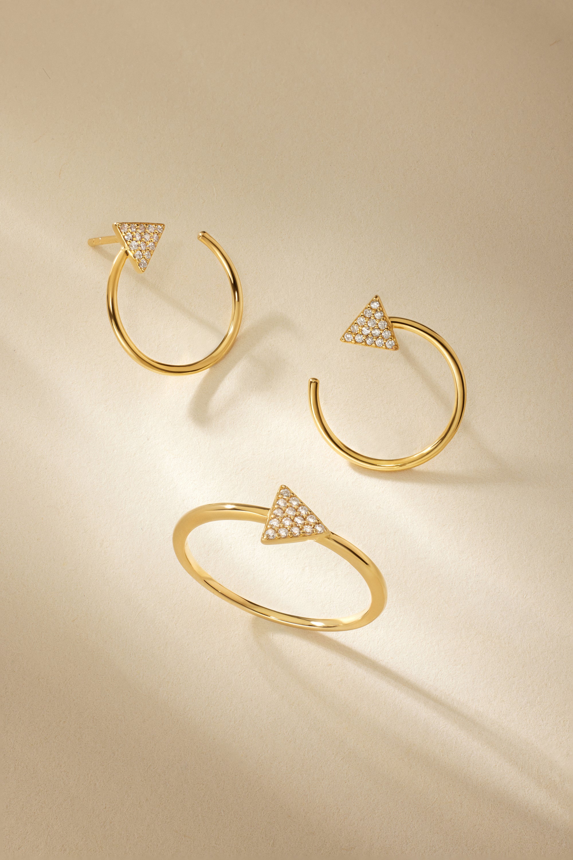 Three Reneé Triangle Linear Hoop Stud Earrings in Gold, each adorned with a diamond-studded triangle, are artfully arranged on a beige background with soft shadows.