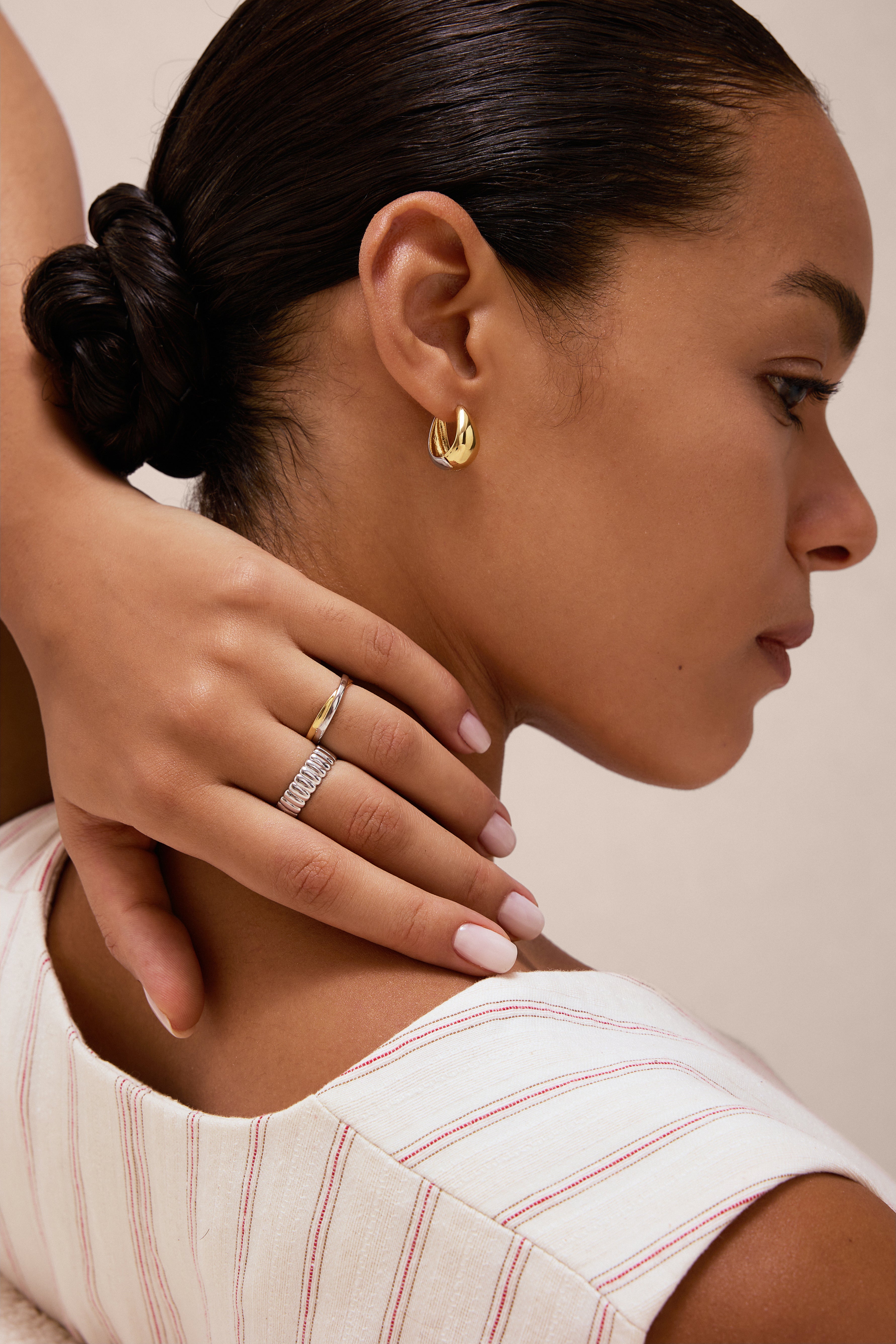 A woman with smooth skin and dark hair in a low bun wears gold hoop earrings and a Charlotte Eternity Ridged Ring in Silver, her hand resting on her neck. She is dressed in a cream top with red stripes and has light pink manicured nails.
