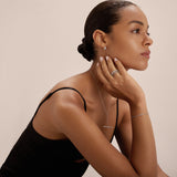 A woman with slicked-back hair wears a black sleeveless top and the Colette Droop Link Huggie Earrings in Silver, along with silver rings, a necklace, and a bracelet. She gazes thoughtfully to the side with her chin resting on her hand.