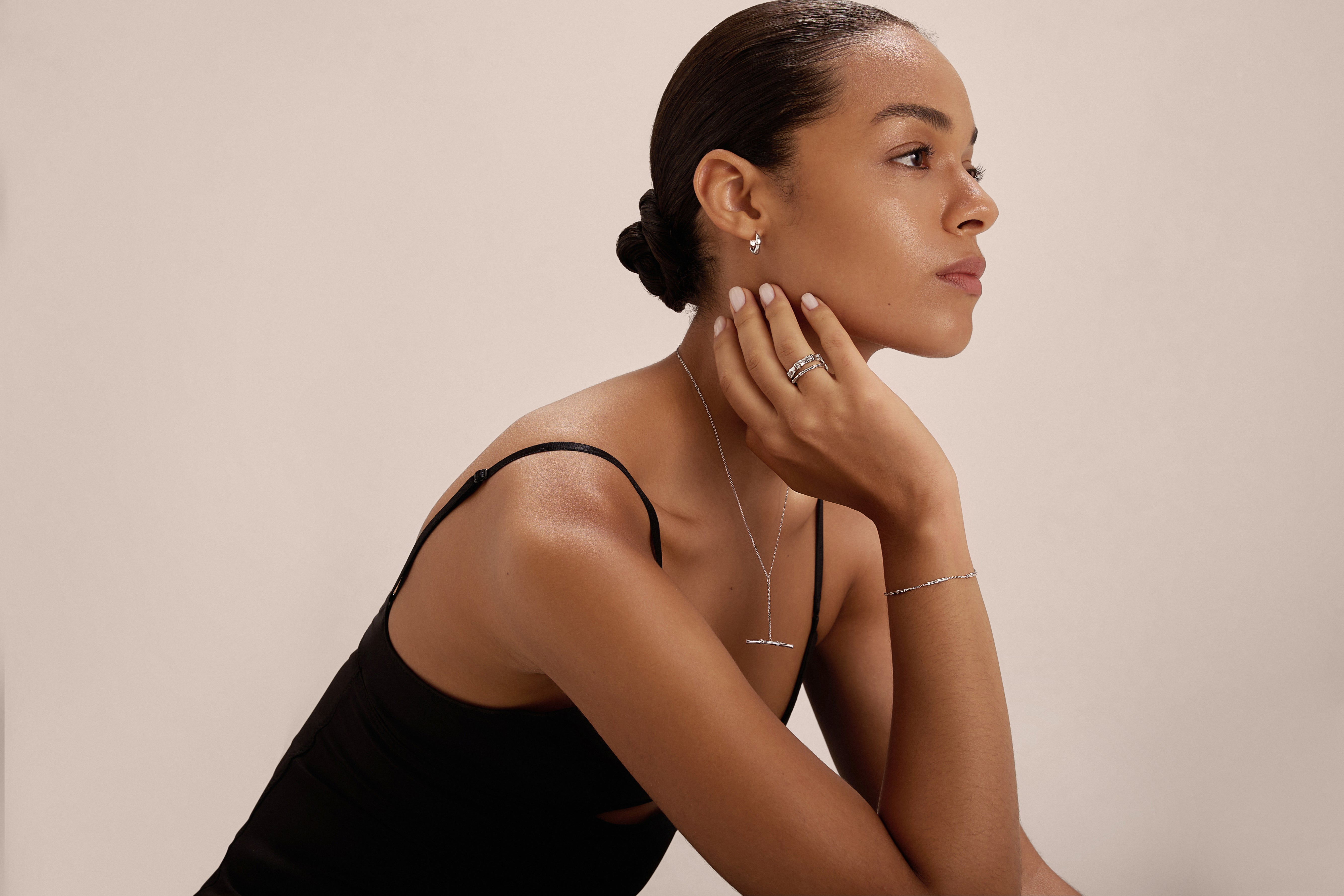 A woman with slicked-back hair wears a black sleeveless top and the Colette Droop Link Huggie Earrings in Silver, along with silver rings, a necklace, and a bracelet. She gazes thoughtfully to the side with her chin resting on her hand.