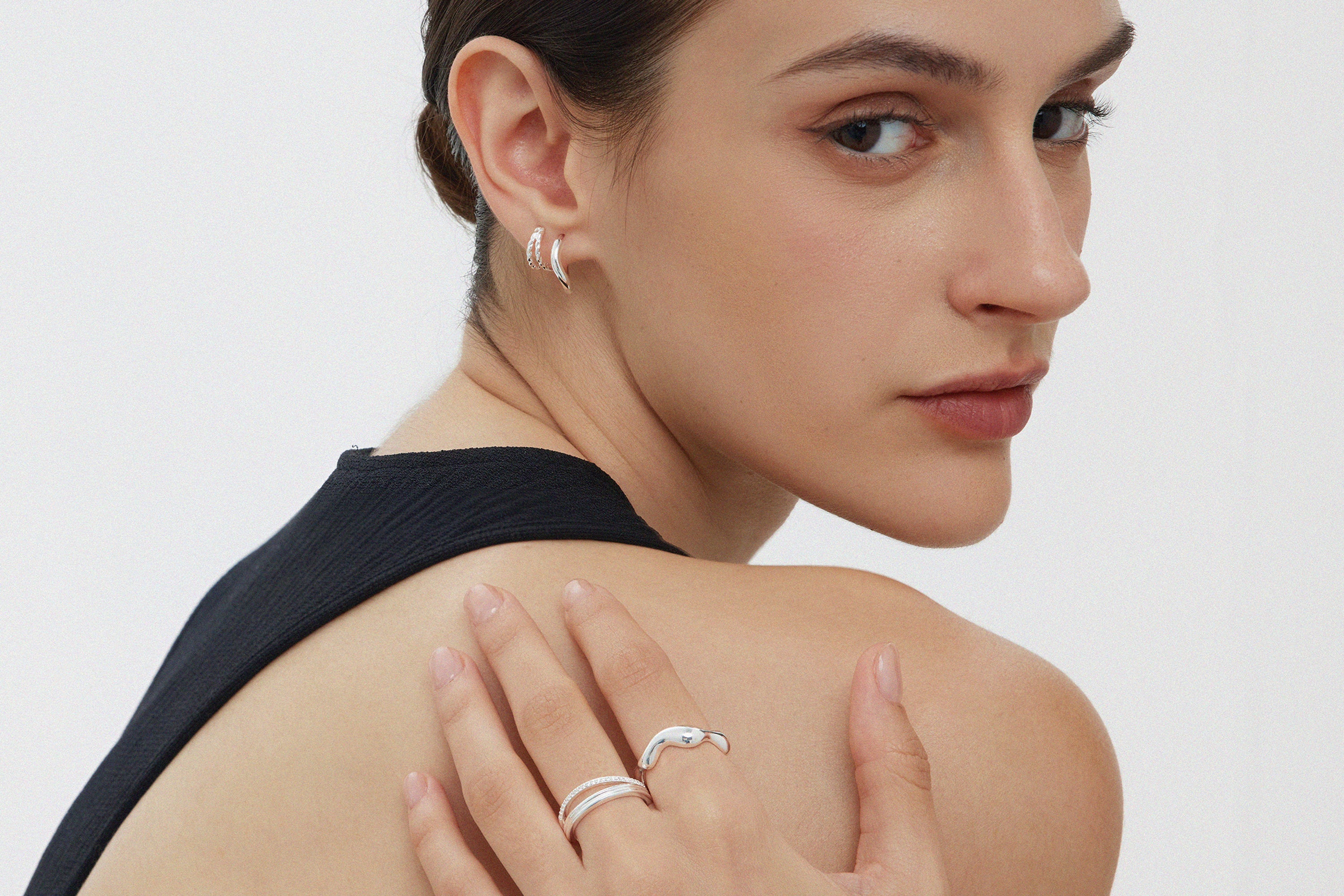 A woman with short, dark hair wears silver hoop earrings and the Vica Fluid Wave Chunky Band Ring in Silver, posing with her hand on her shoulder. She is dressed in a sleeveless black top against a plain white background.