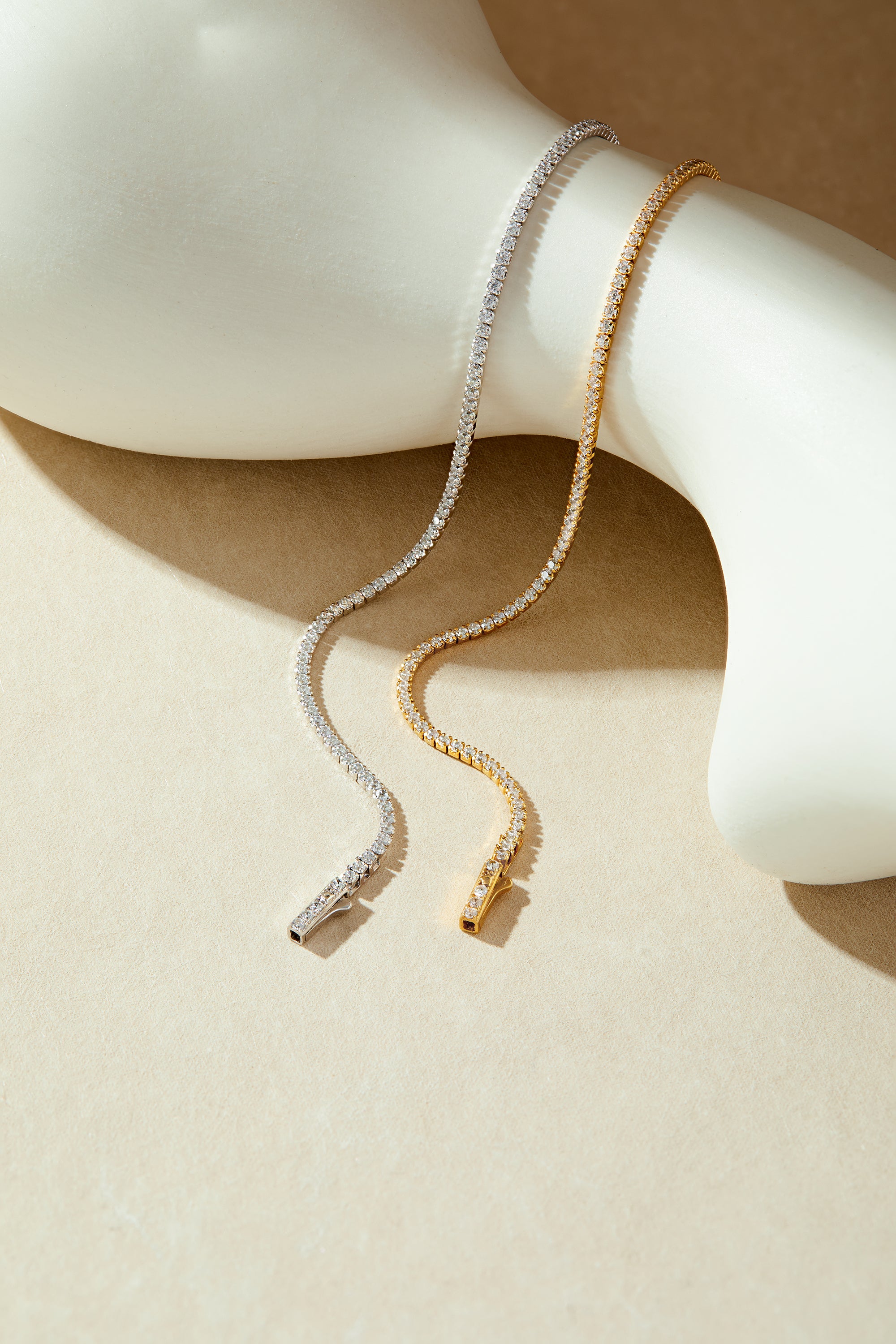 The Alba Tennis Necklace in Gold, adorned with sparkling cubic zirconia, drapes elegantly over a smooth, white abstract sculpture on a beige background.