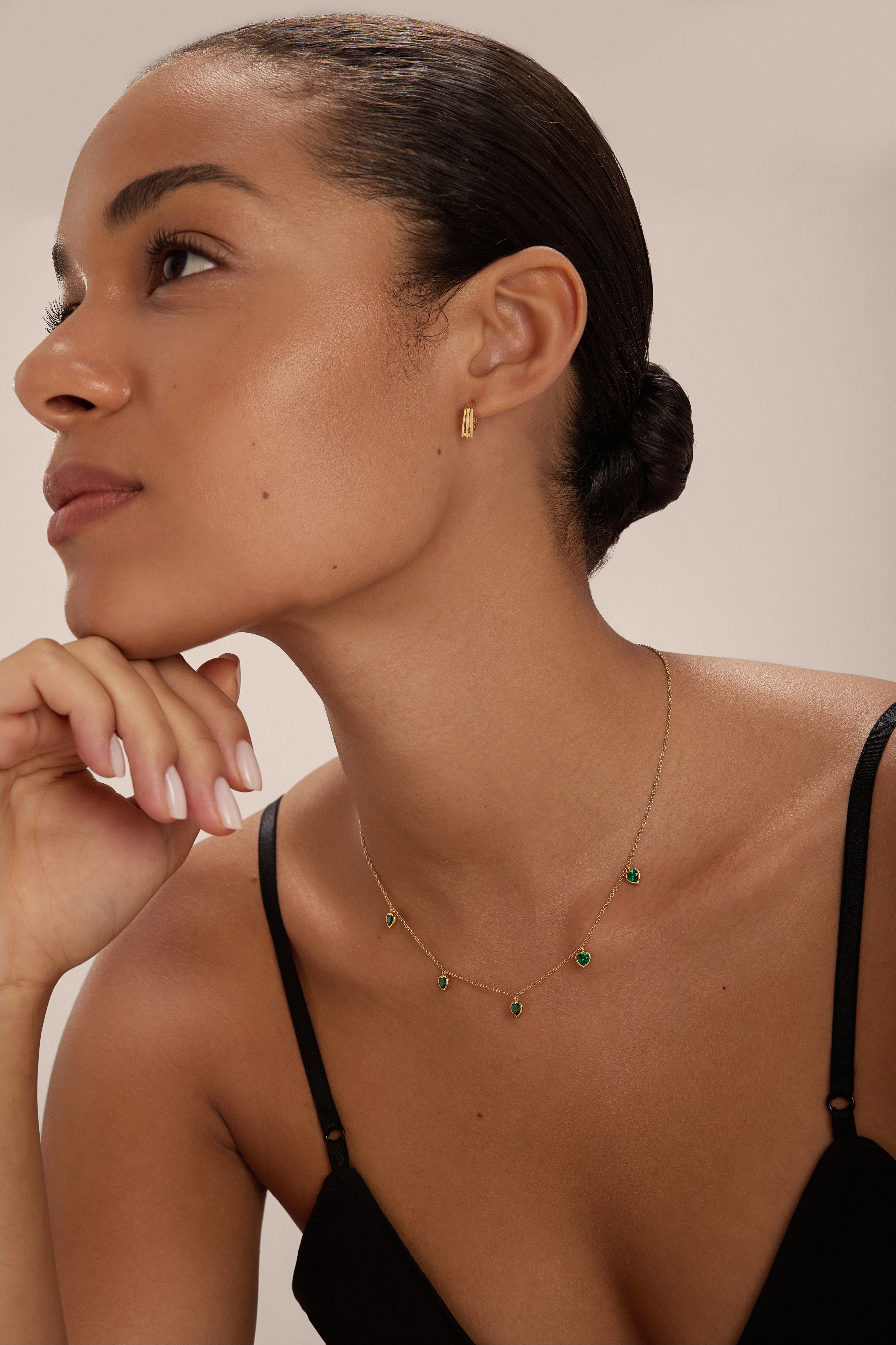 A woman with smooth, glowing skin and dark hair tied back wears the Thea Green Heart Pendant Gold Necklace with matching gold earrings. She poses in a black spaghetti strap top against a neutral background.