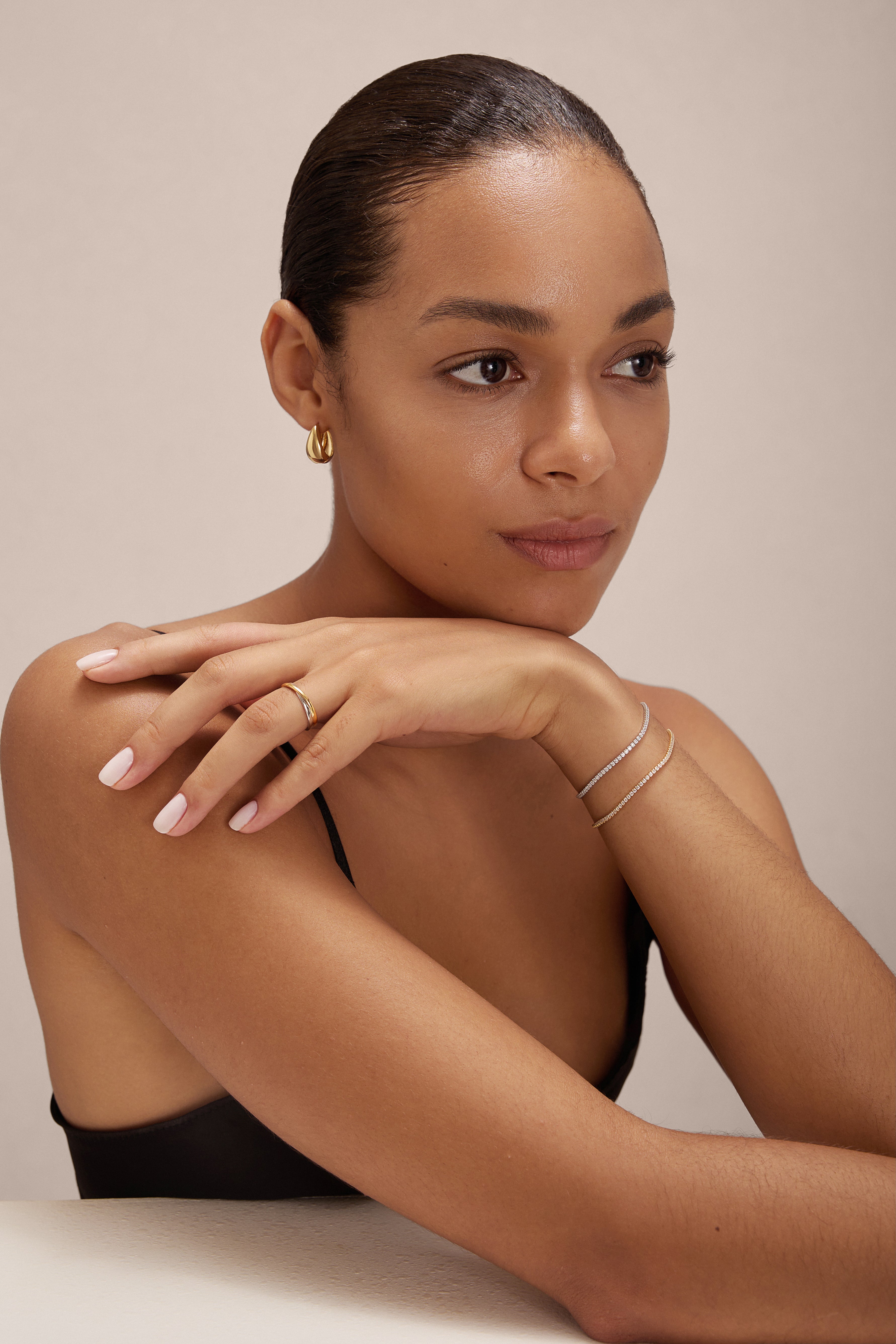 A woman with smooth, medium skin and slicked-back hair rests her chin on her hand, wearing gold hoop earrings, a thin bracelet, the Yvette Two-Tone Mixed Metal Ring and another ring, plus a black top, natural makeup and light pink nails.