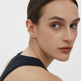 A woman with slicked-back dark hair and natural makeup wears a sleeveless black top, glancing over her shoulder. She sports the Vale Crystal Ear Cuff in Gold on her left ear, adding shimmer and sparkle against a plain white background.