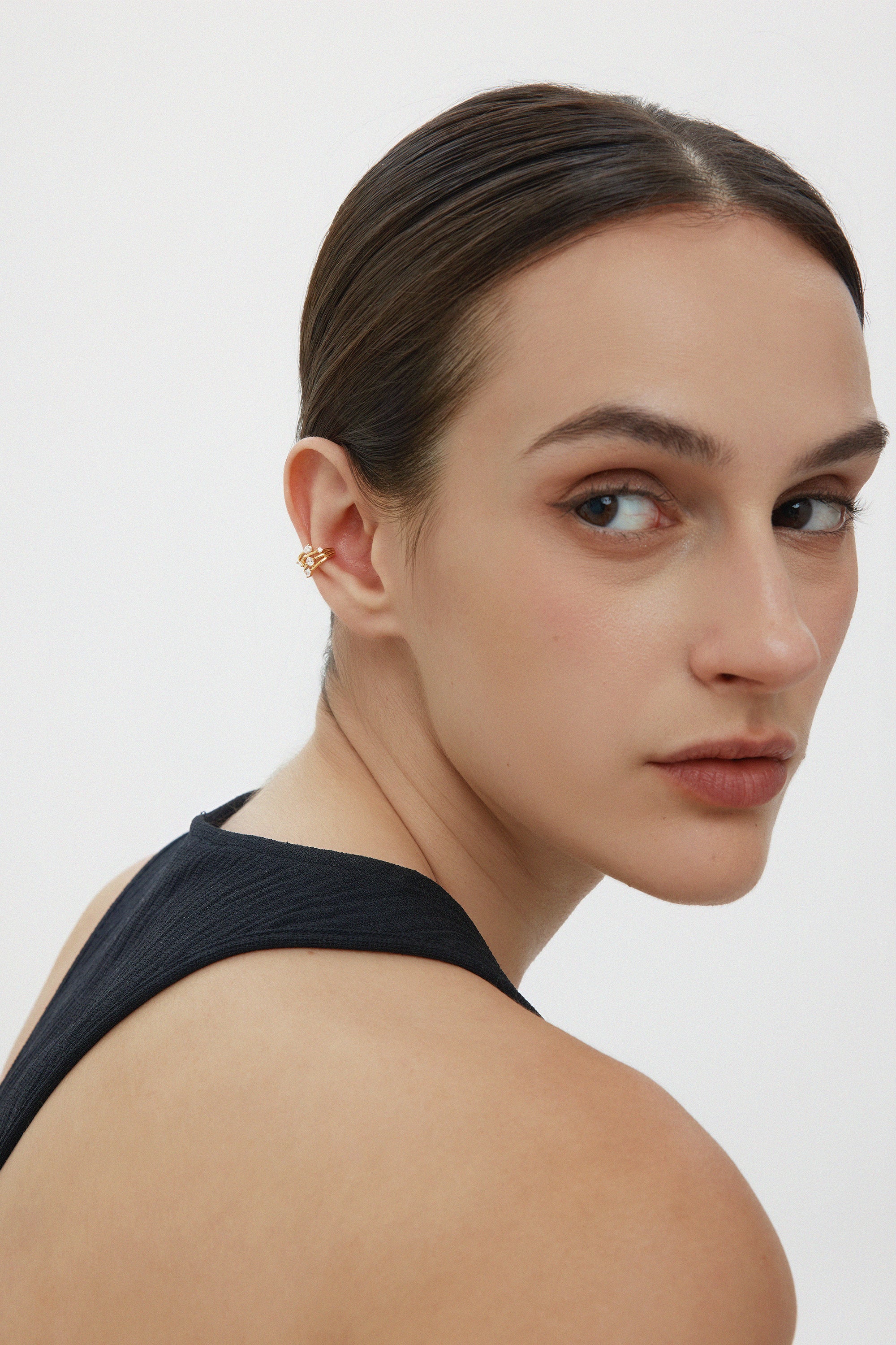 A woman with slicked-back dark hair and natural makeup wears a sleeveless black top, glancing over her shoulder. She sports the Vale Crystal Ear Cuff in Gold on her left ear, adding shimmer and sparkle against a plain white background.