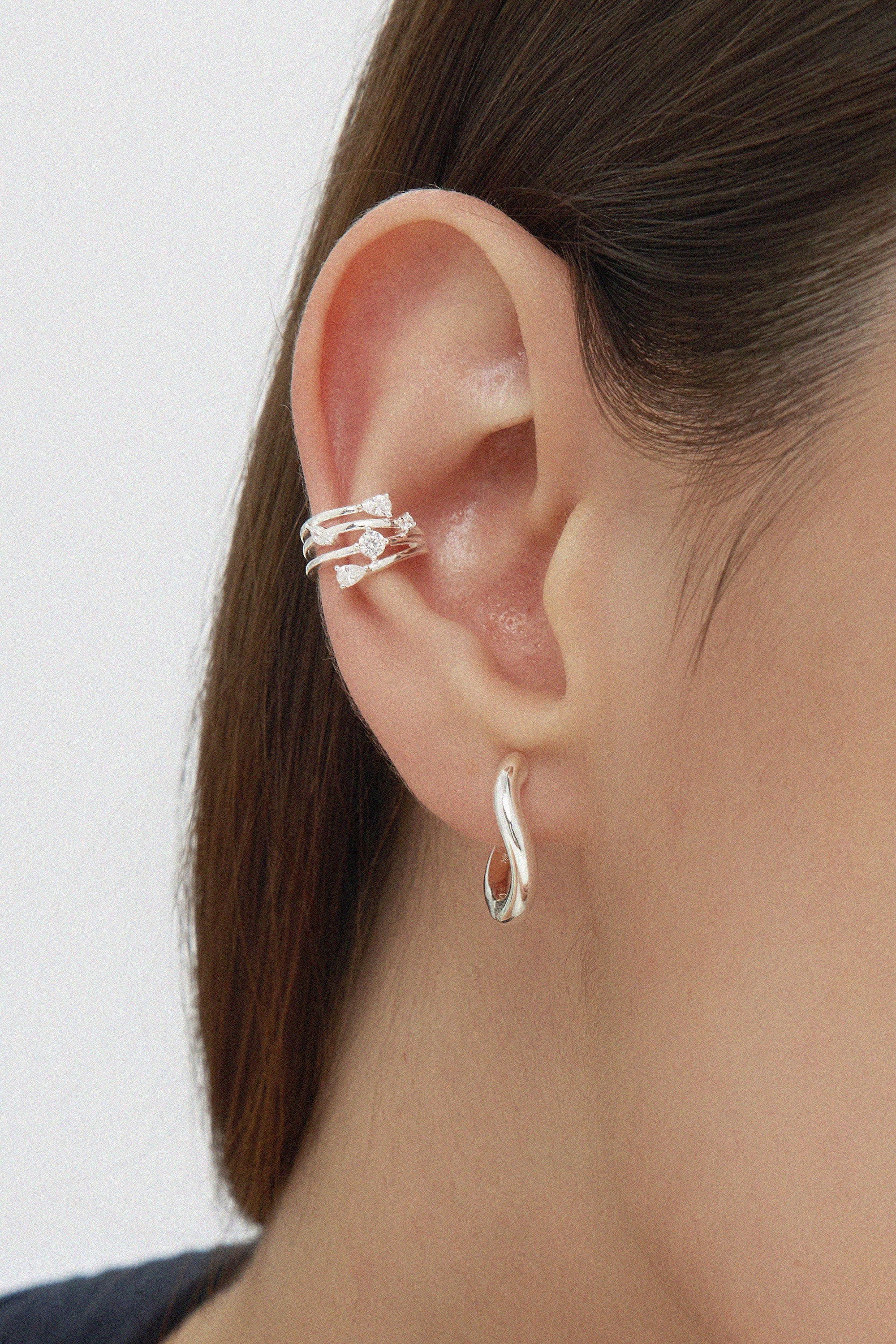 A close-up shows brown hair tucked behind an ear styled with an elegant stack: the Vale Crystal Ear Cuff in Silver featuring sparkling gemstones on the upper ear and a twisted silver hoop earring in the lobe.