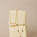 The Thea Green Heart Pendant Gold Necklace, featuring a small green cubic zirconia heart, is draped over two stacked blocks of butter against a neutral beige background.