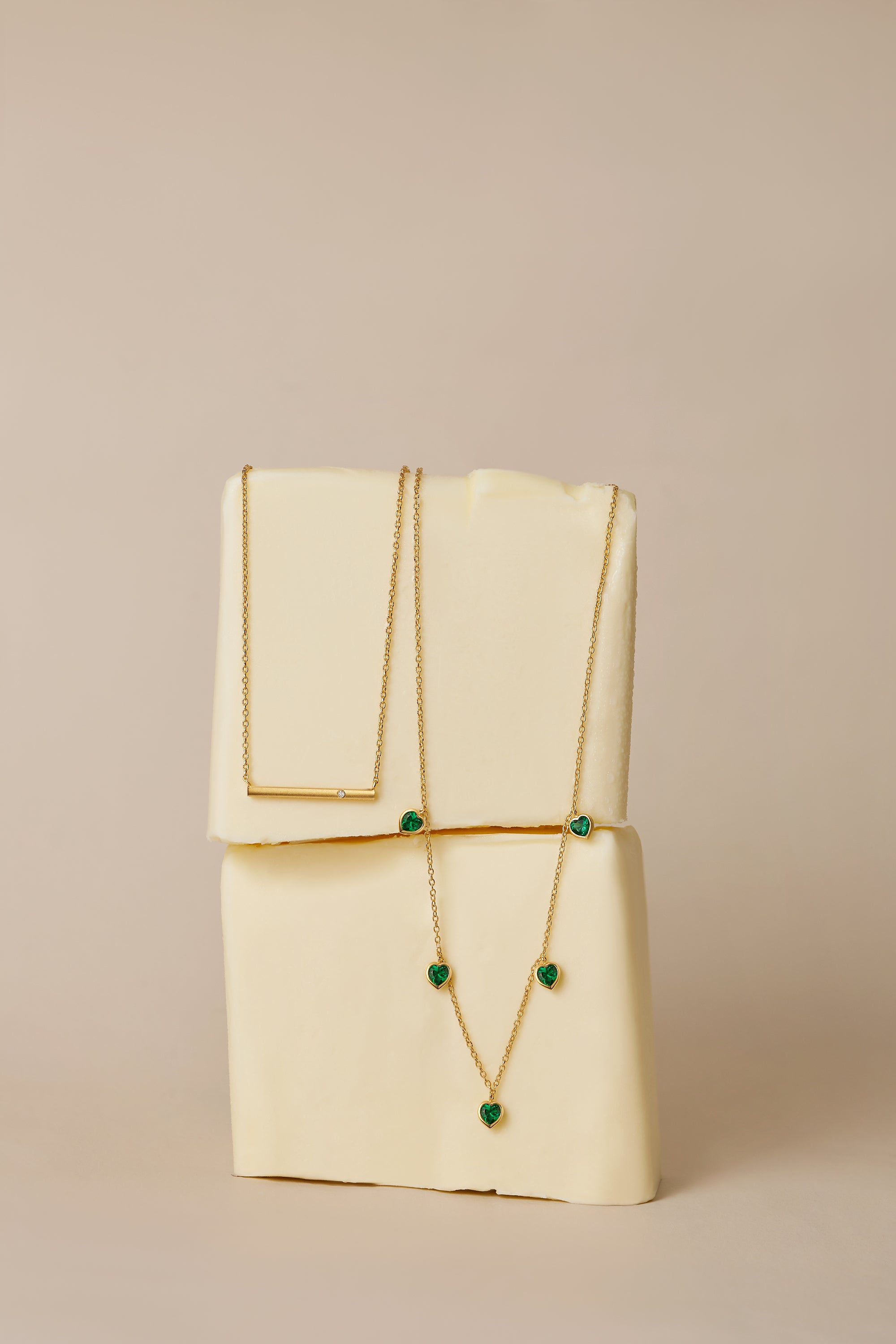 The Thea Green Heart Pendant Gold Necklace, featuring a small green cubic zirconia heart, is draped over two stacked blocks of butter against a neutral beige background.