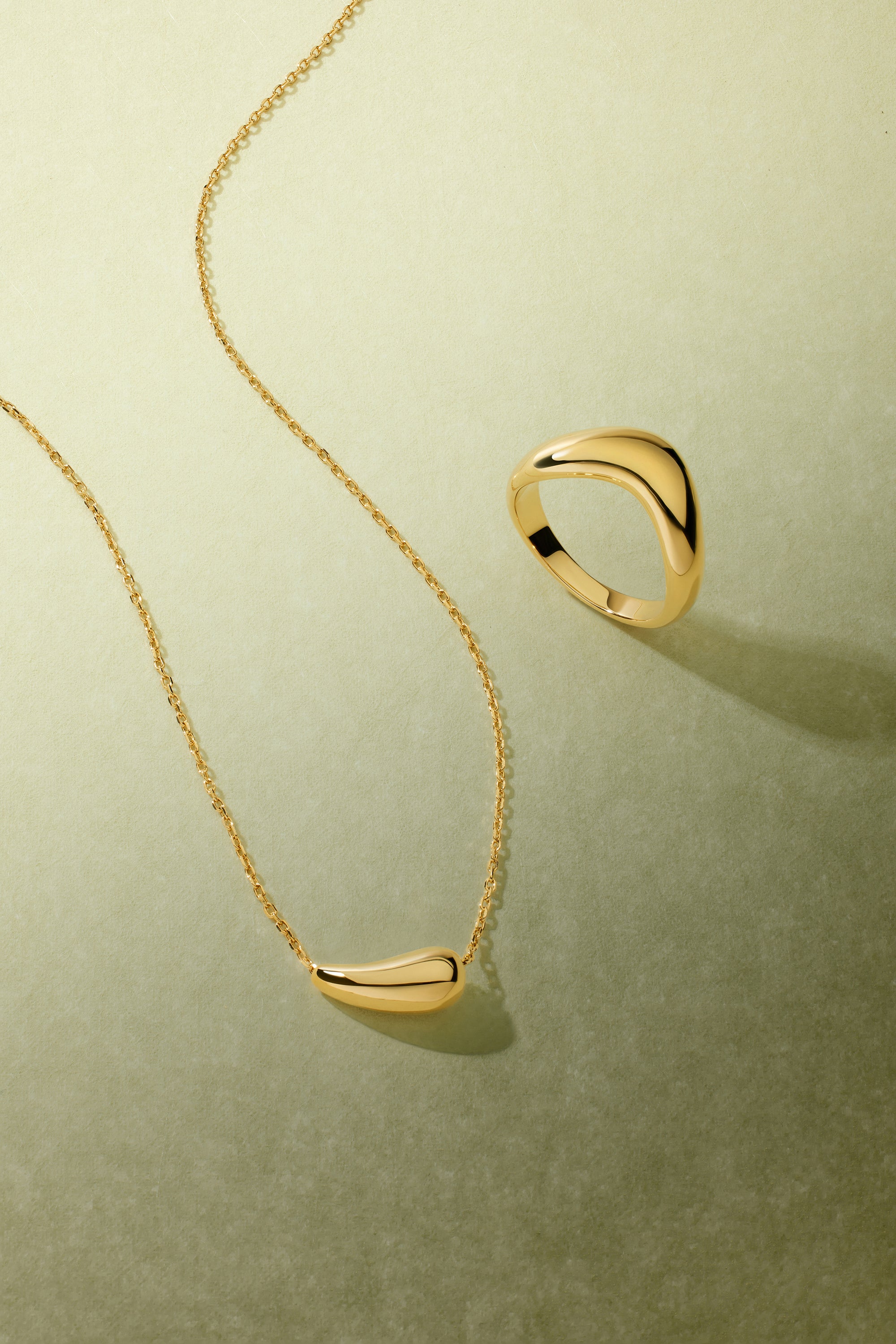 Displayed on a light green textured surface, the Vava Plain Chunky Curved Wave Ring in Gold and a matching abstract necklace cast soft shadows—a striking minimalist jewelry duo.
