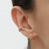 A close-up of a person’s ear wearing two gold hoop earrings and the Vera Twisted Crystal Ear Cuff in Gold, all against a plain light background.