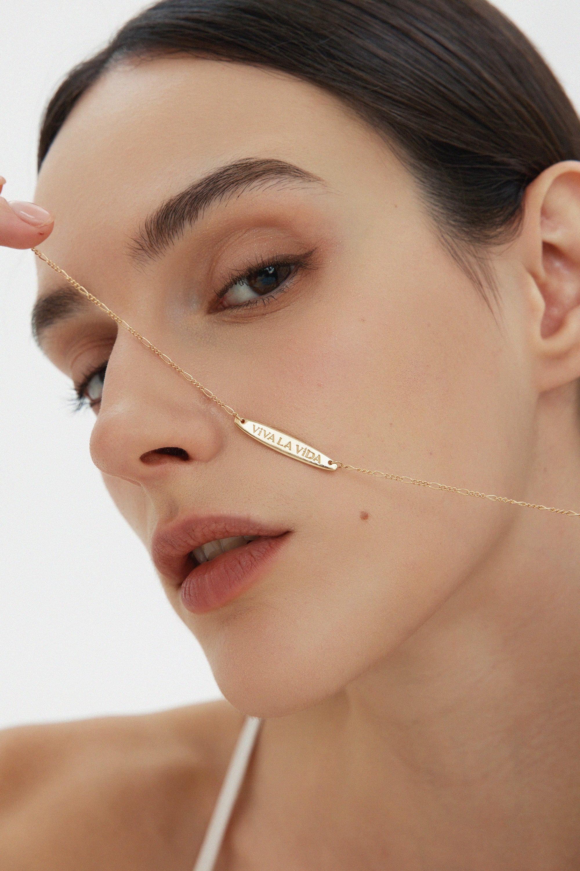 A woman with smooth skin holds the Viva La Vida Bar Chain Necklace in Gold, its adjustable figaro chain glinting as she gazes neutrally into the camera against a simple, light backdrop.