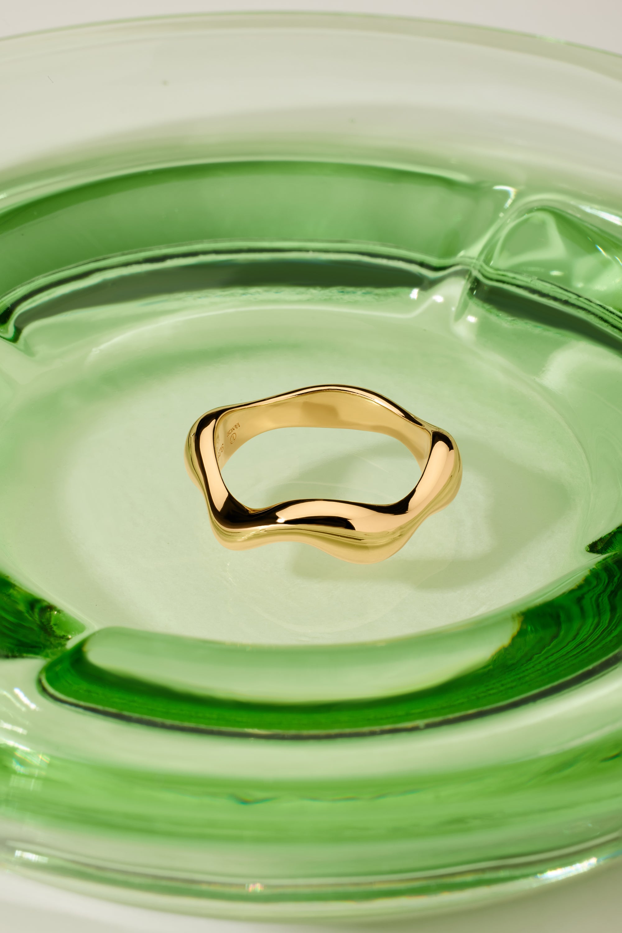 The Vica Fluid Wave Chunky Band Ring in Gold sits at the center of a smooth, modern clear green glass dish.