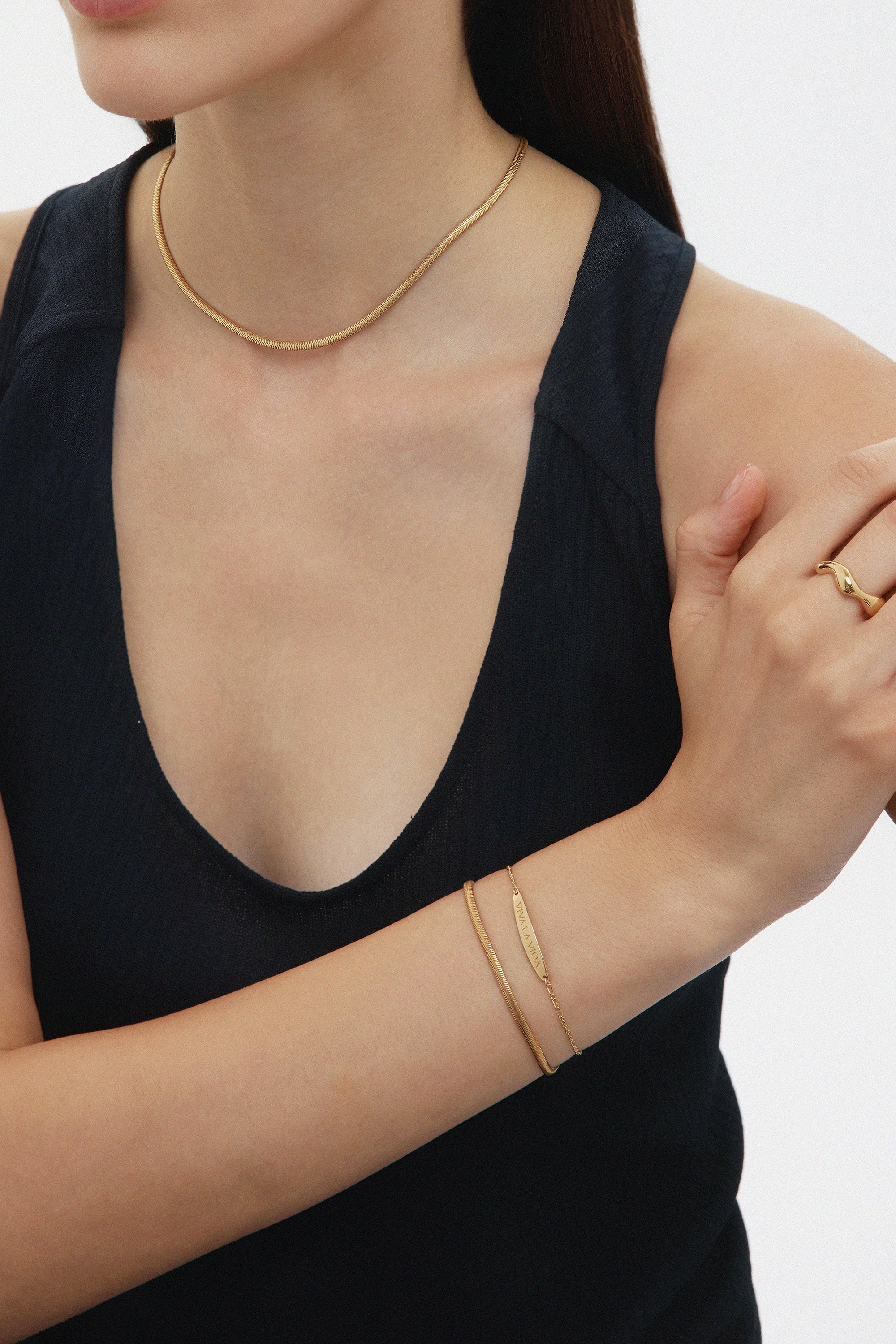 A woman in a sleeveless black top wears minimal gold jewelry—including the Vica Fluid Wave Chunky Band Ring in Gold—with her arms crossed and face partially out of frame against a plain background.