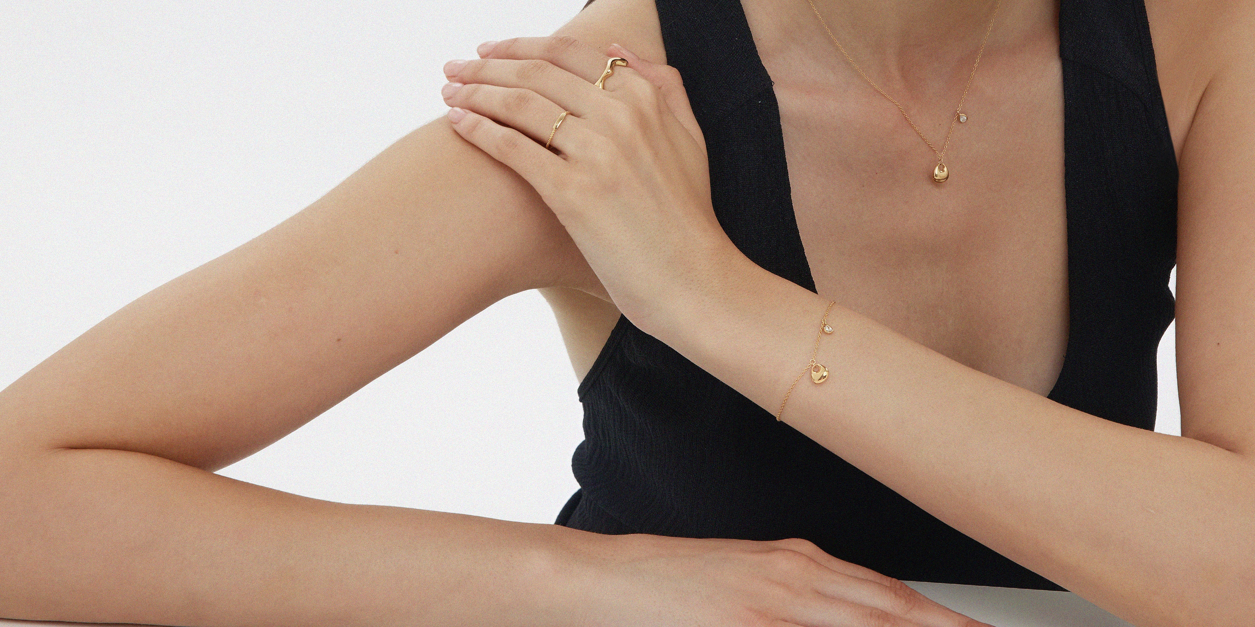 A person in a sleeveless black top rests an arm on a table, wearing the Vica Fluid Wave Chunky Band Ring in Gold along with other delicate gold jewelry. They pose against a plain white background.