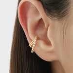 Close-up of a persons ear wearing the Vina Crystal Bezel Ear Cuff in Gold, adorned with small round cubic zirconia. Straight brown hair frames the ear, set against a softly blurred background.