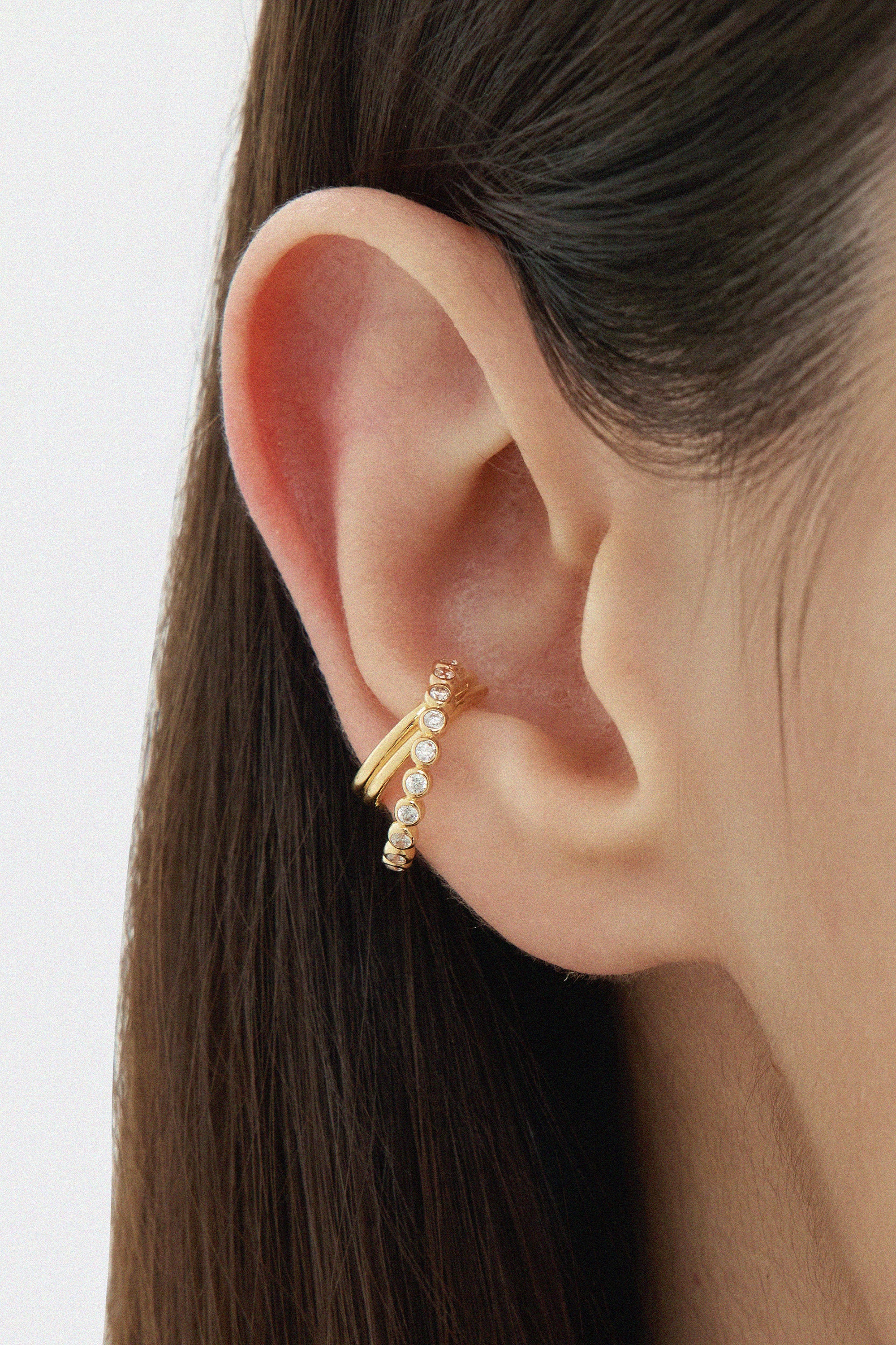 Close-up of a persons ear wearing the Vina Crystal Bezel Ear Cuff in Gold, adorned with small round cubic zirconia. Straight brown hair frames the ear, set against a softly blurred background.
