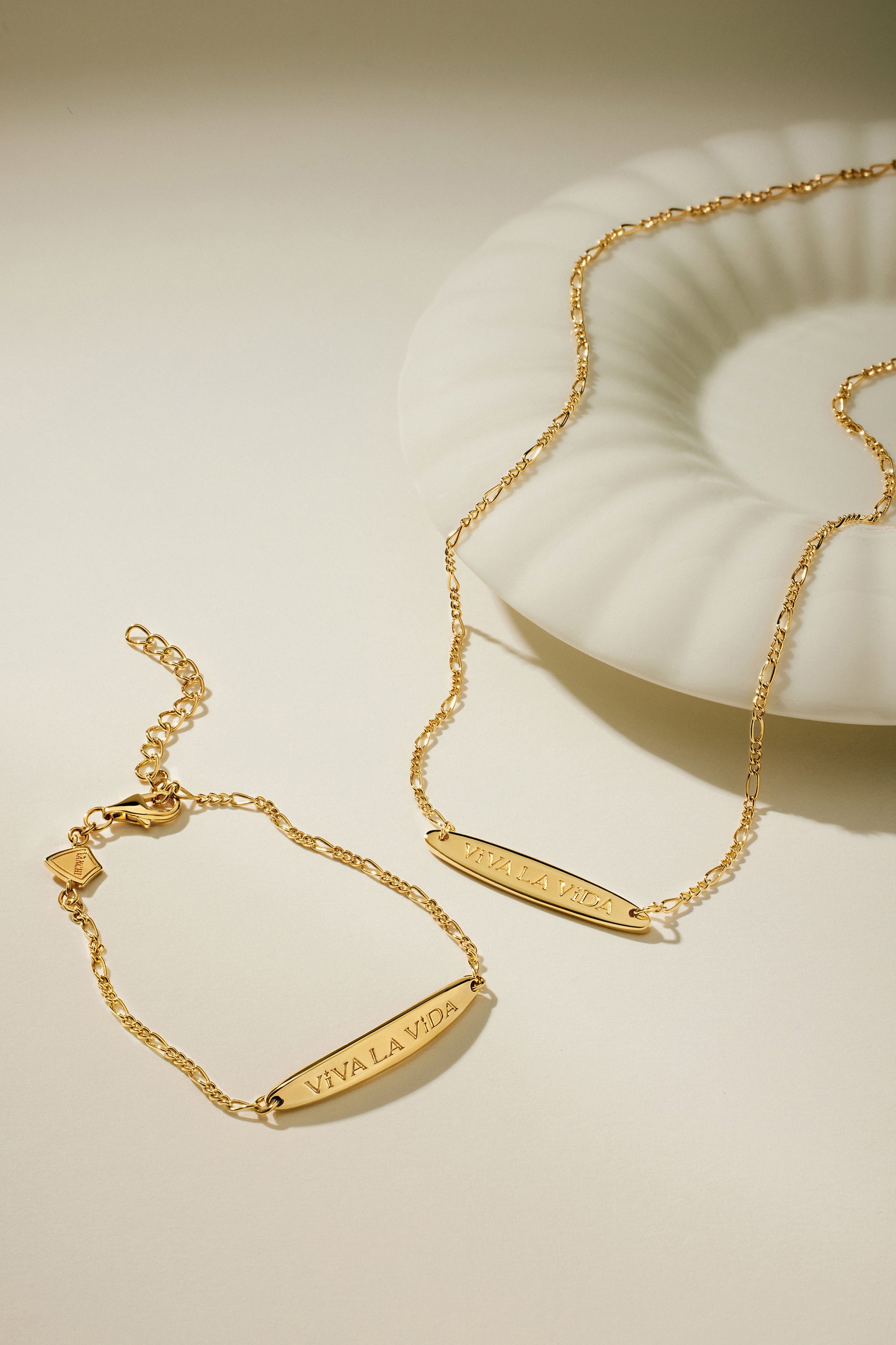 The Viva La Vida Bar Chain Bracelet in Gold, featuring an engraved bar and adjustable figaro chain, is displayed beside a white shell-shaped dish on a neutral background.
