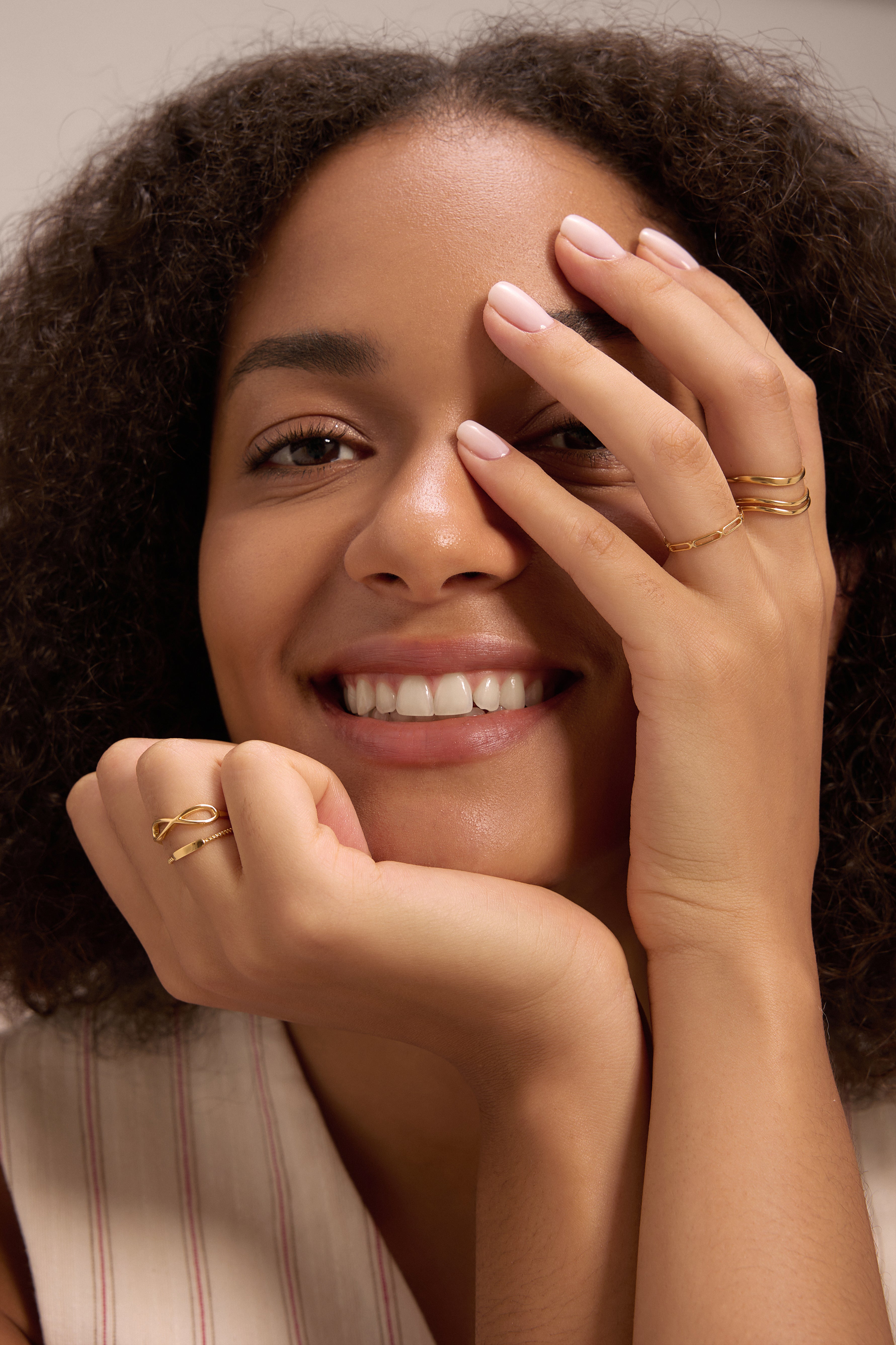 A smiling woman with curly hair rests her chin on one hand and covers one eye with the other, wearing the Zoe Triple Wave Ring in Gold and light pink nail polish.