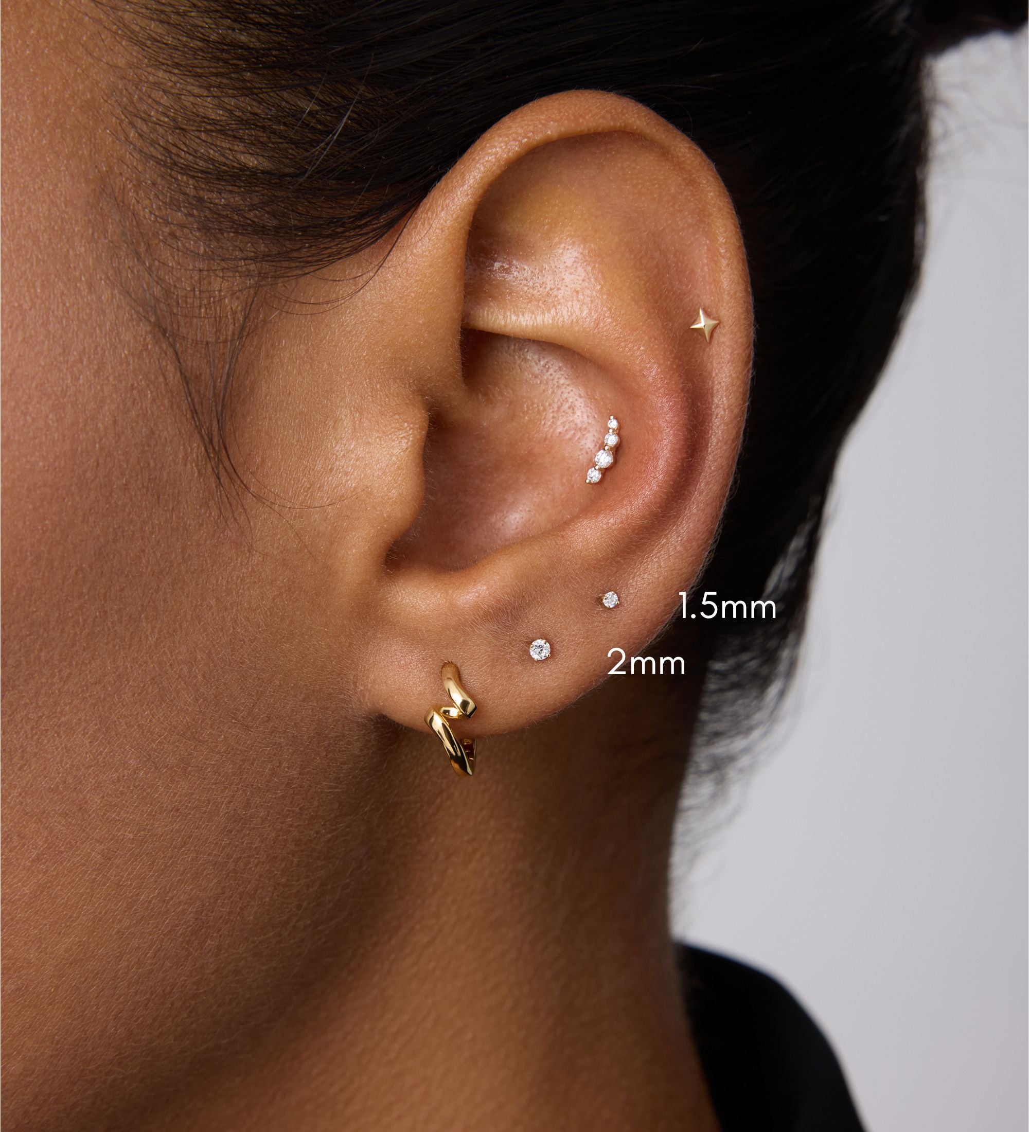 A close-up of a person’s ear with multiple piercings, including a 14K Solid White Gold Lab-grown Diamond Prong Set Flat Back Stud (1.5mm), plus gold and silver studs and a gold hoop. The person has medium brown skin and dark hair.