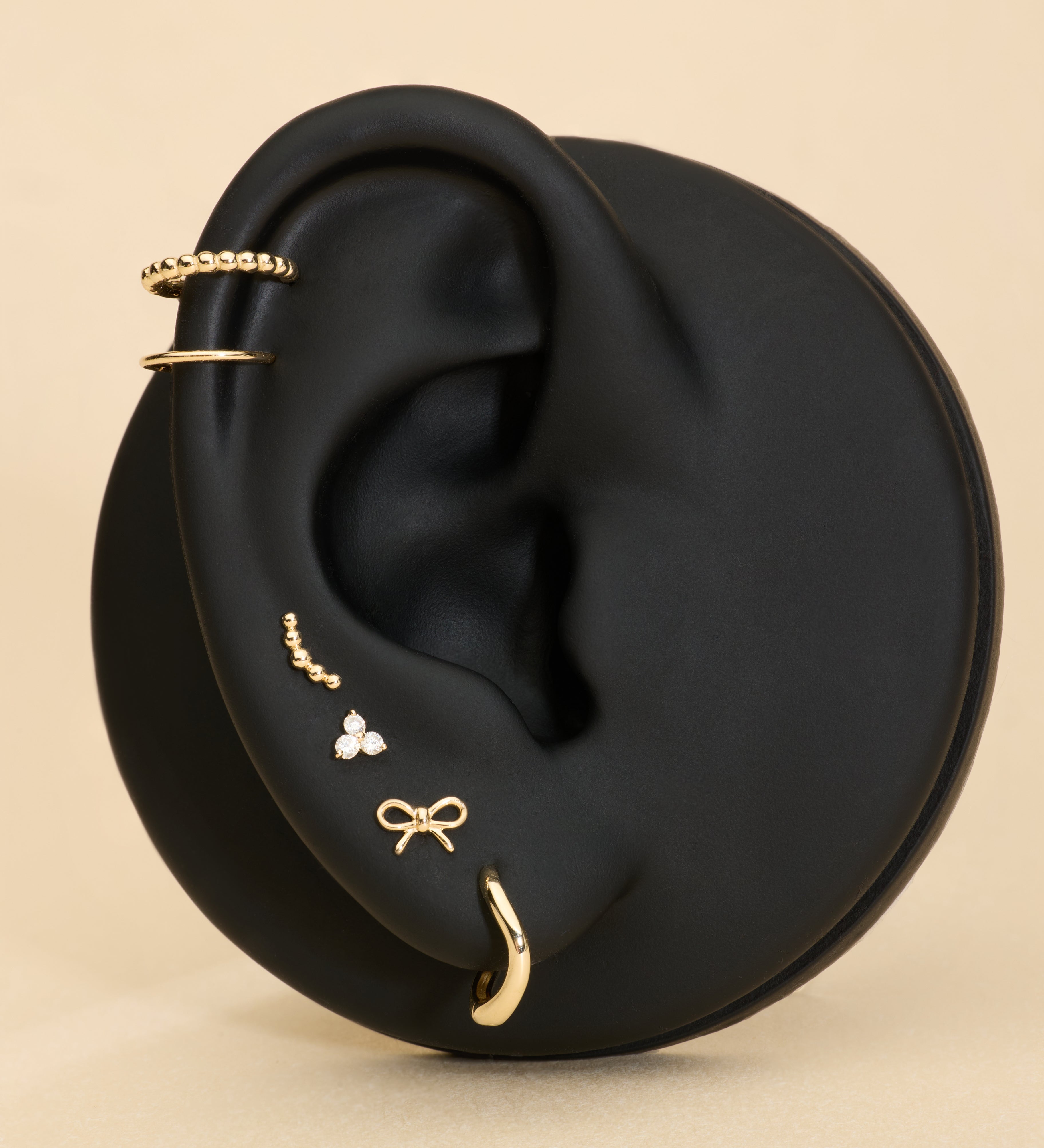 A black mannequin ear displays 14K Recycled Solid Yellow Gold Twisted Huggie Earrings among other gold styles against a beige background.