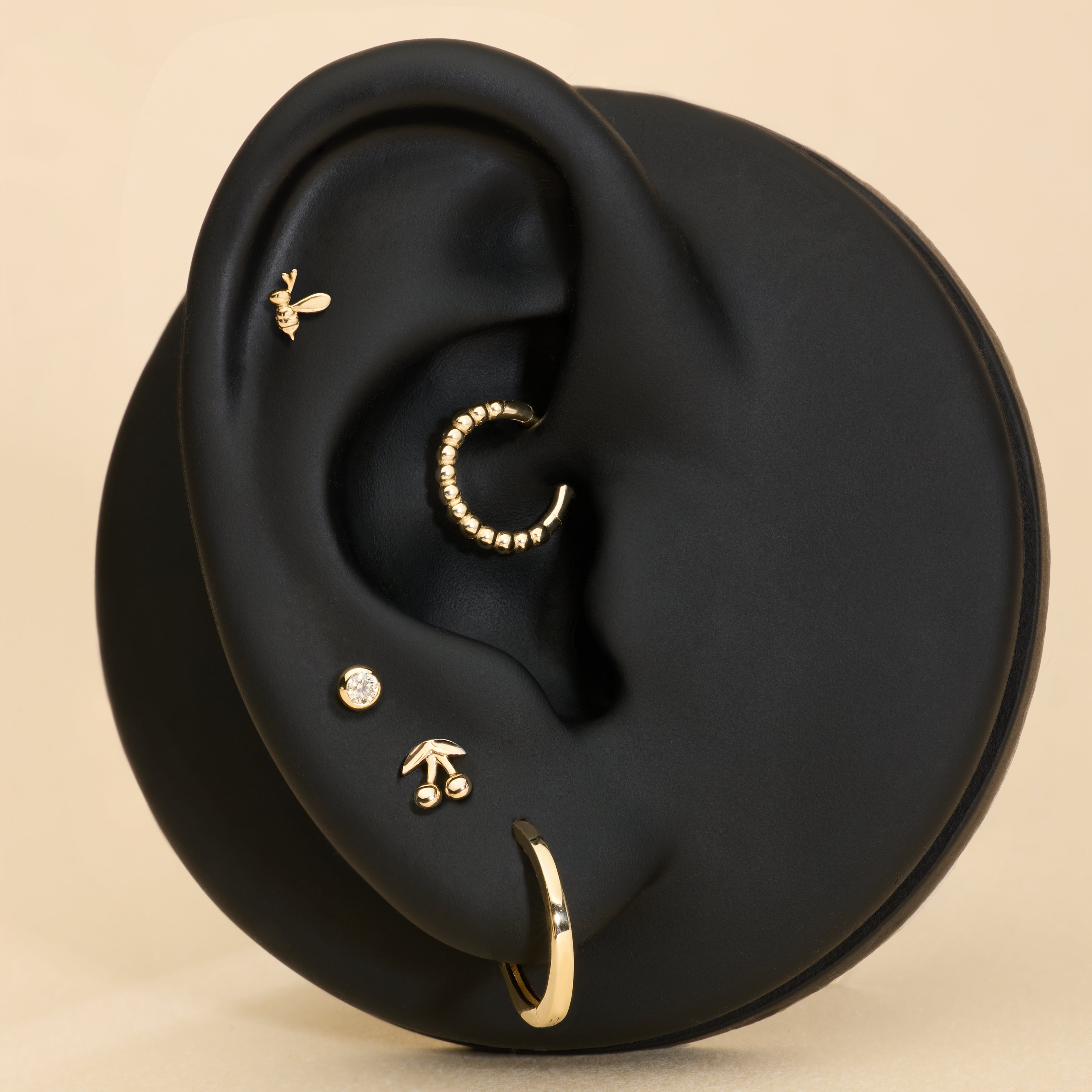 A black ear model displays five gold earrings, including the 14K Solid White Gold Bumble Bee Flat Back Stud (Left), with a beige background.
