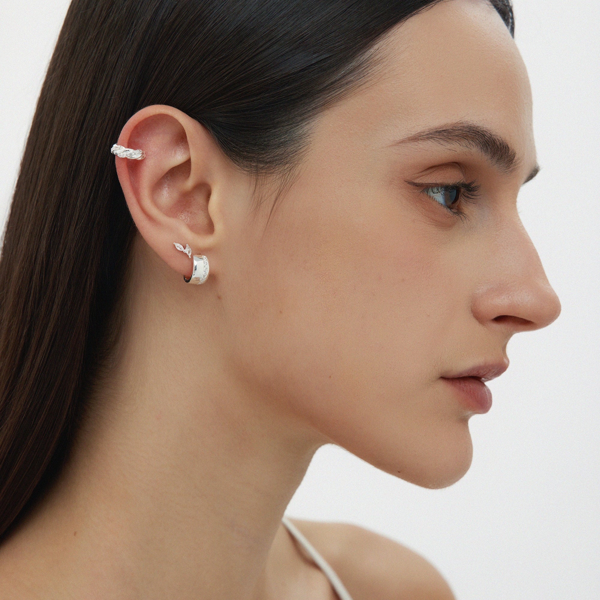 A woman with straight dark hair and a white top is shown in profile, wearing the Flora Barbell Stud in Gold adorned with marquise cubic zirconia on her upper ear and a textured silver hoop on her lobe, set against a plain light background.