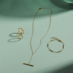 A gold necklace, ring, and Bamboo Chain Bracelet in Gold with delicate, minimalist designs and bamboo-inspired details are arranged on a light green surface, casting soft shadows in the sunlight.