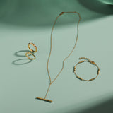 A gold necklace, ring, and Bamboo Chain Bracelet in Gold with delicate, minimalist designs and bamboo-inspired details are arranged on a light green surface, casting soft shadows in the sunlight.