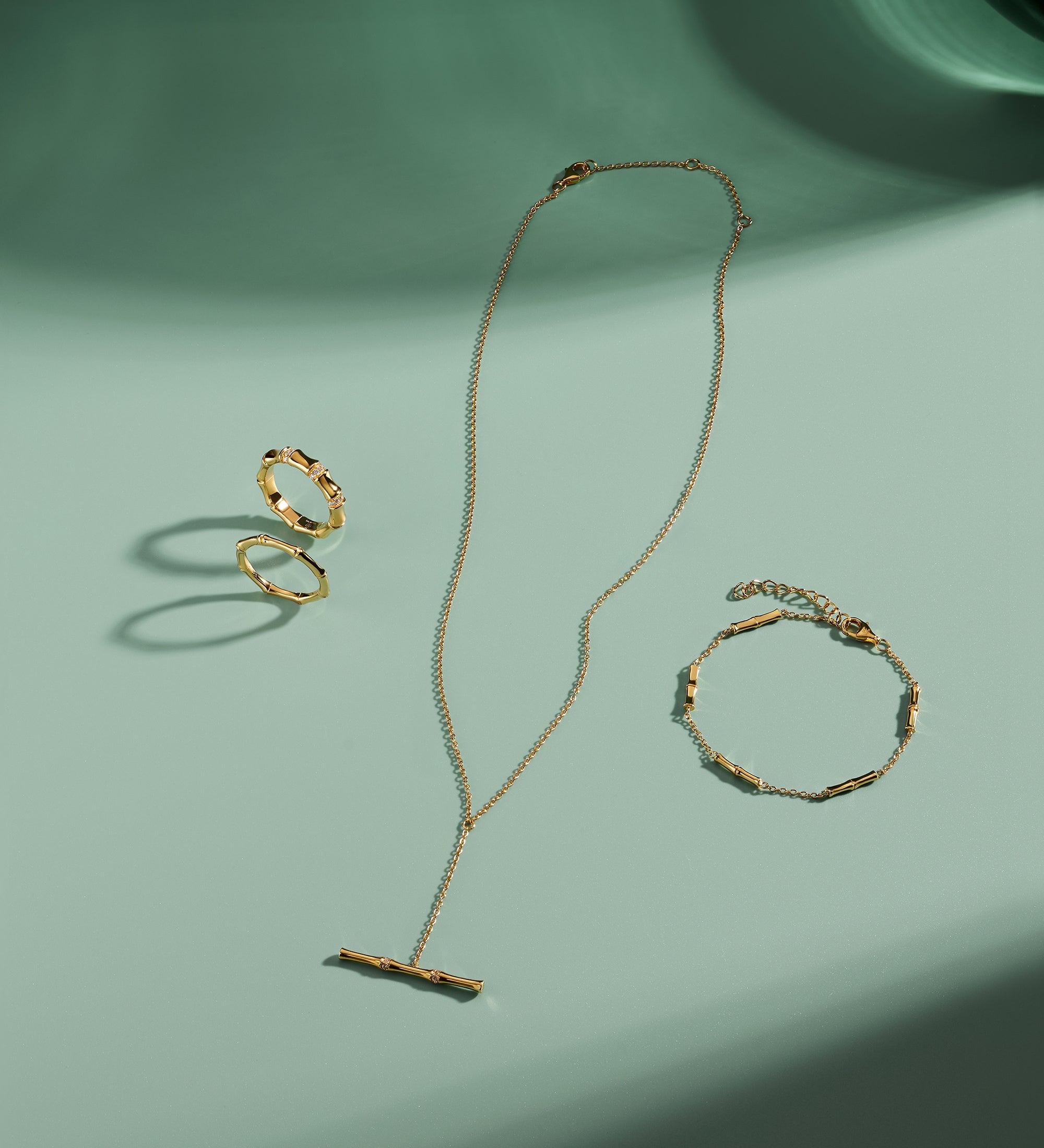 A gold necklace, ring, and Bamboo Chain Bracelet in Gold with delicate, minimalist designs and bamboo-inspired details are arranged on a light green surface, casting soft shadows in the sunlight.