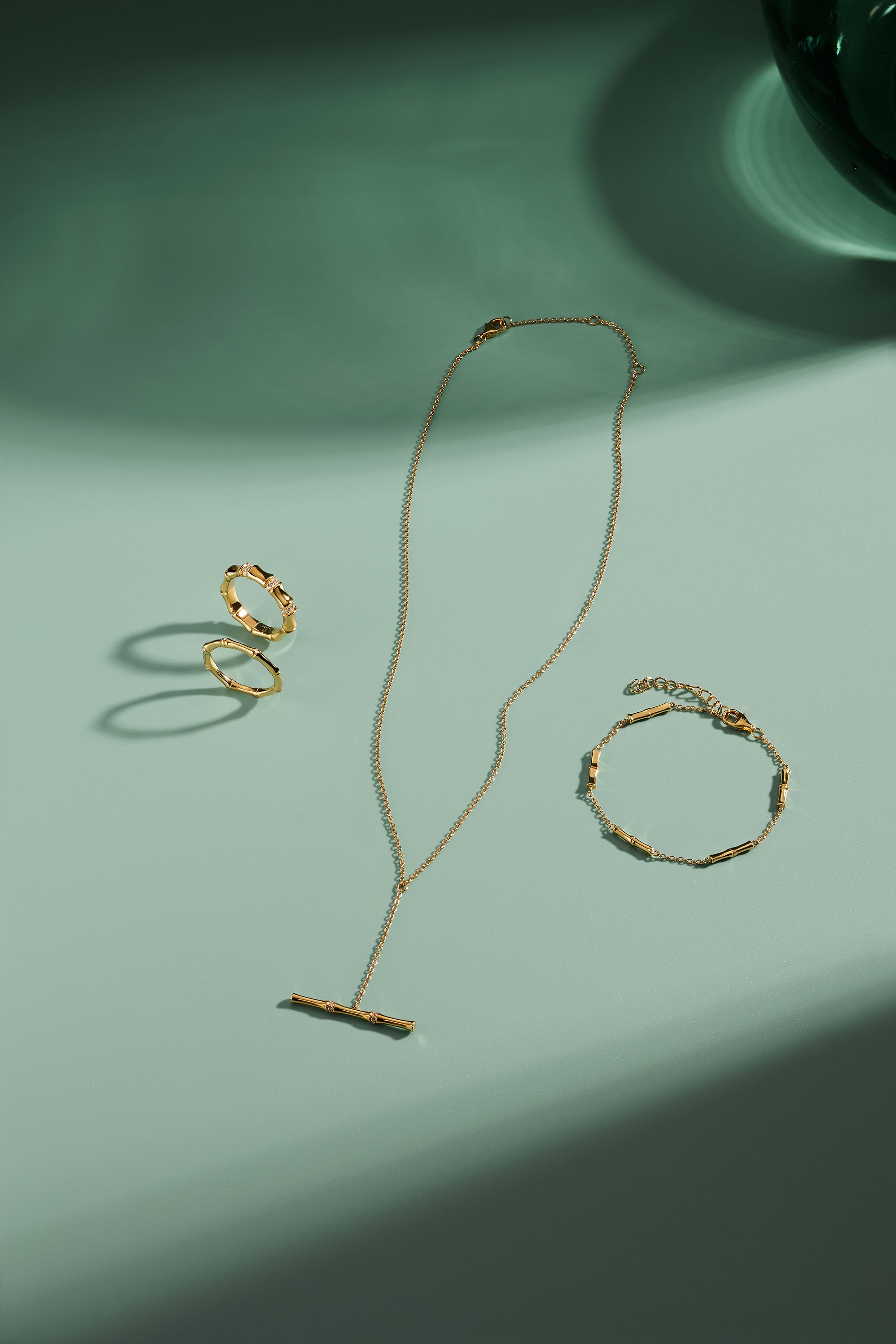 A gold necklace, ring, and Bamboo Chain Bracelet in Gold with delicate, minimalist designs and bamboo-inspired details are arranged on a light green surface, casting soft shadows in the sunlight.