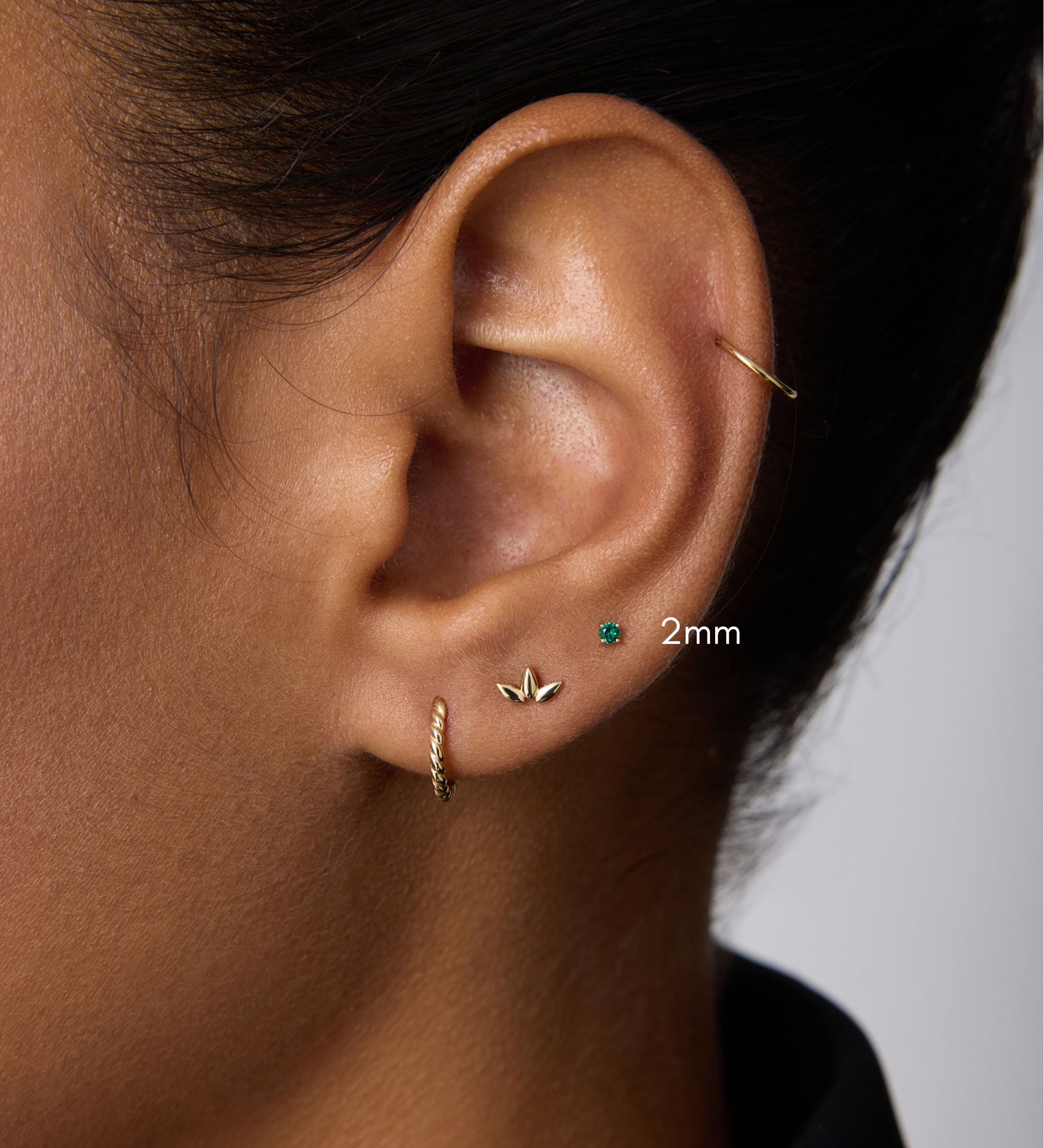 A close-up of an ear with four earrings: a 14k gold small hoop, a leaf-shaped stud, the 14K Solid Yellow Gold Green Emerald Flat Back Stud - 2mm, and a thin hoop on the upper ear. The person wears a black collared shirt.