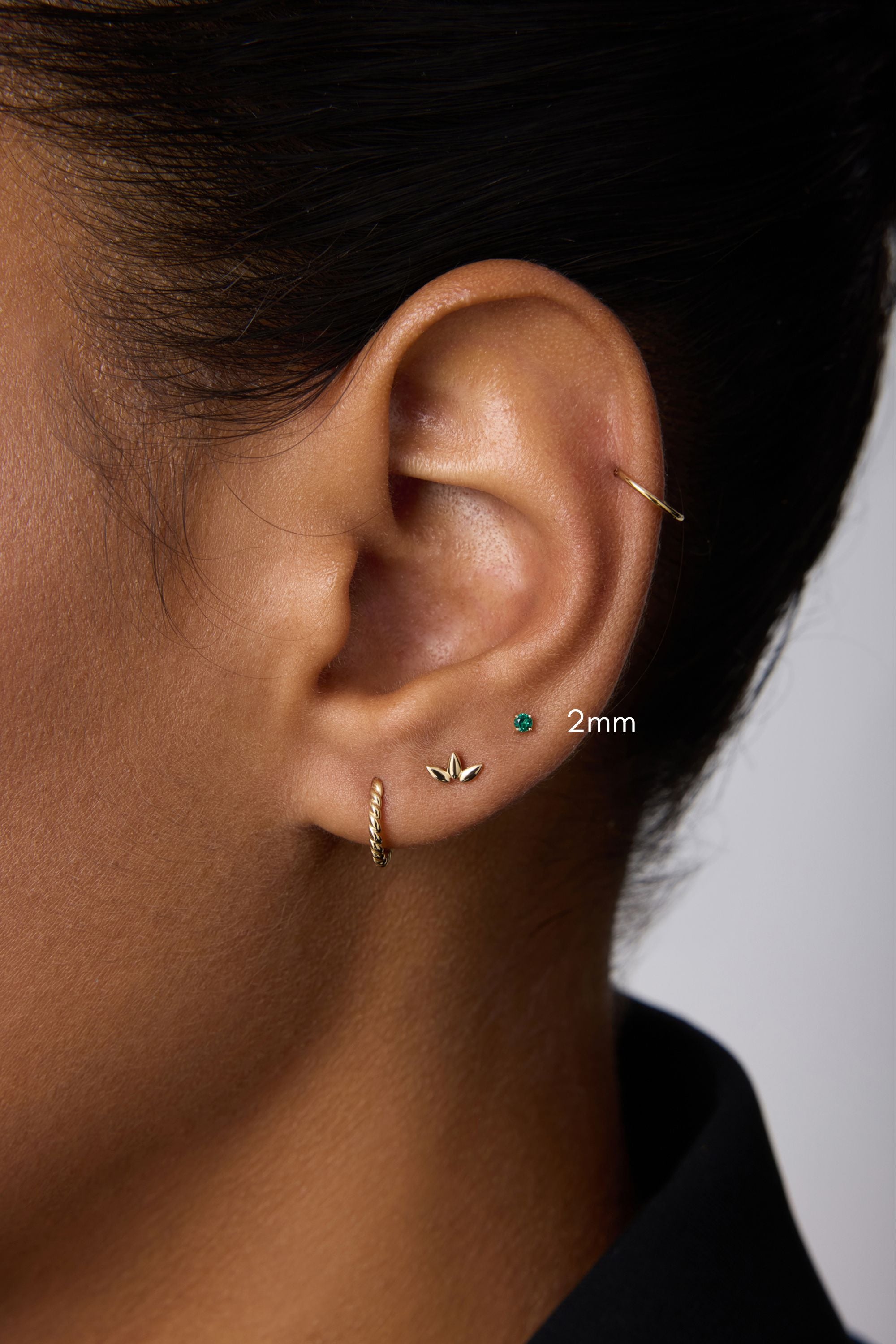 A close-up of an ear with four earrings: a 14k gold small hoop, a leaf-shaped stud, the 14K Solid Yellow Gold Green Emerald Flat Back Stud - 2mm, and a thin hoop on the upper ear. The person wears a black collared shirt.