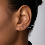 A close-up of a person’s ear with four piercings, featuring three small 14k gold stud earrings—1.5mm and a 2mm Flat Back Stud (14K Solid Yellow Gold Green Emerald)—plus a small hoop earring. The background is plain; the person wears a dark top.