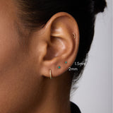 A close-up of a person’s ear with four piercings, featuring three small 14k gold stud earrings—1.5mm and a 2mm Flat Back Stud (14K Solid Yellow Gold Green Emerald)—plus a small hoop earring. The background is plain; the person wears a dark top.