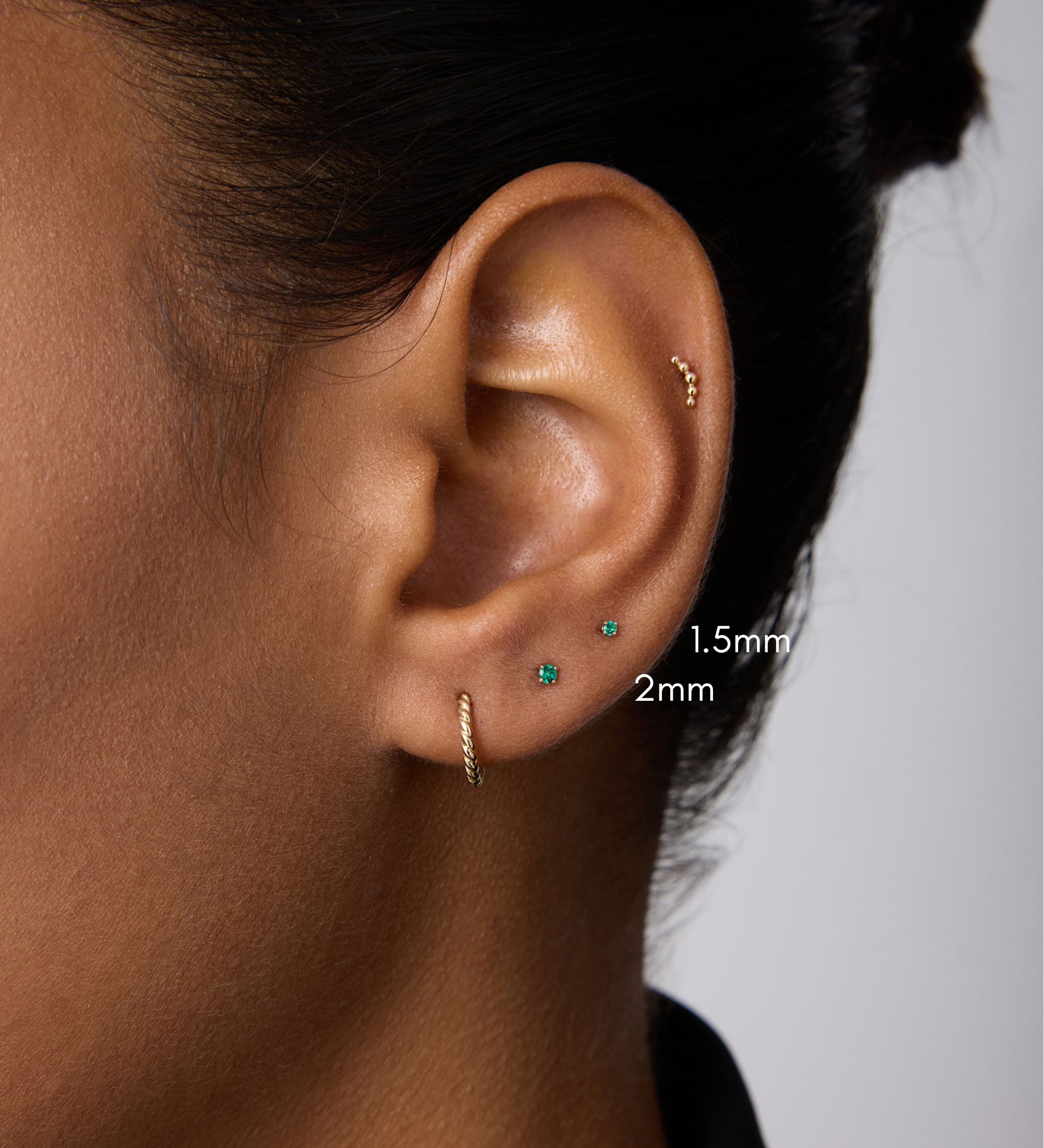 A close-up of a person’s ear with four piercings, featuring three small 14k gold stud earrings—1.5mm and a 2mm Flat Back Stud (14K Solid Yellow Gold Green Emerald)—plus a small hoop earring. The background is plain; the person wears a dark top.