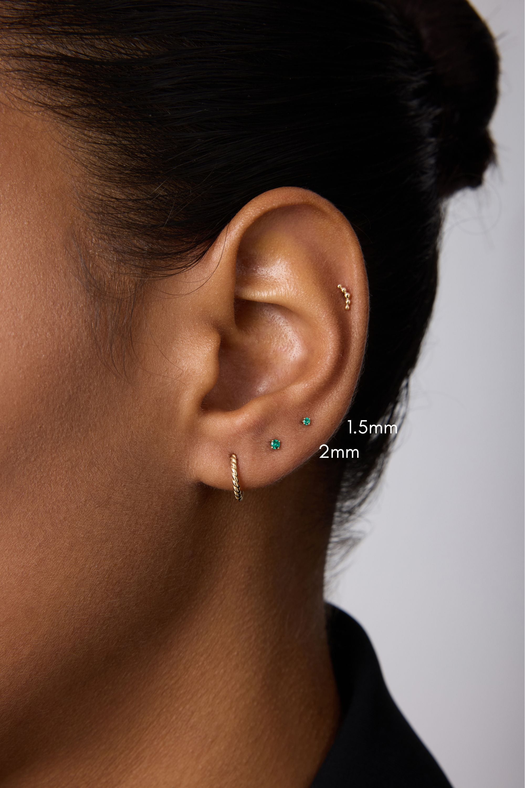 A close-up of a person’s ear with four piercings, featuring three small 14k gold stud earrings—1.5mm and a 2mm Flat Back Stud (14K Solid Yellow Gold Green Emerald)—plus a small hoop earring. The background is plain; the person wears a dark top.