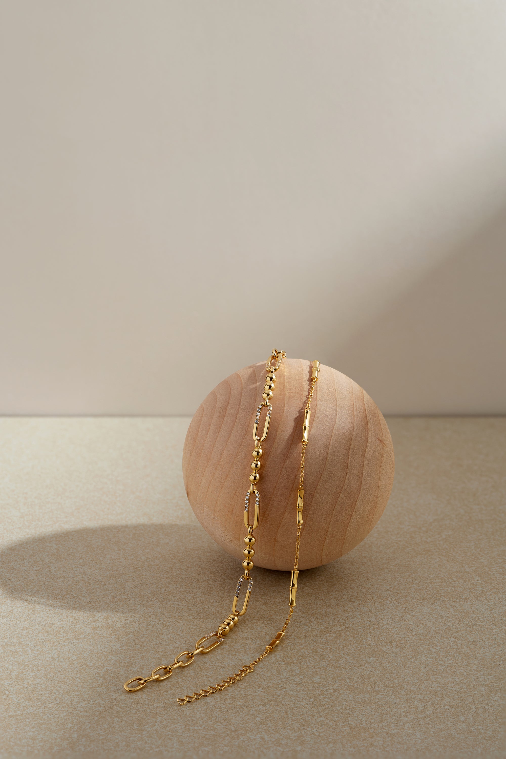 The Bamboo Chain Bracelet in Gold, featuring a delicate 18k gold-plated design, is elegantly displayed draped over a smooth, light wooden sphere against a neutral beige background with soft lighting and gentle shadows.