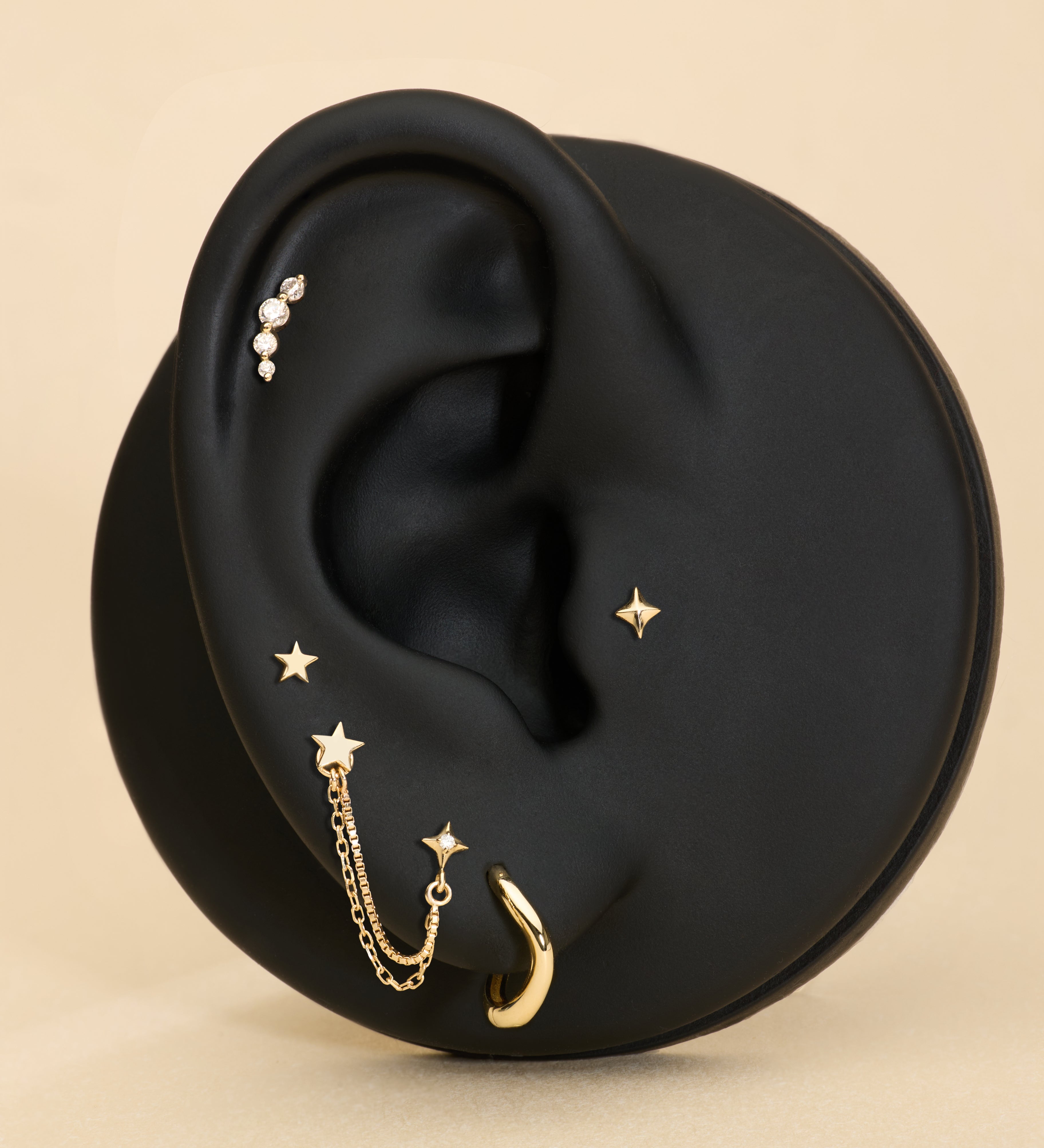 A black ear-shaped display showcases the 14K Solid Yellow Gold Four Point Star Flat Back Stud with other jewelry pieces against a beige background.