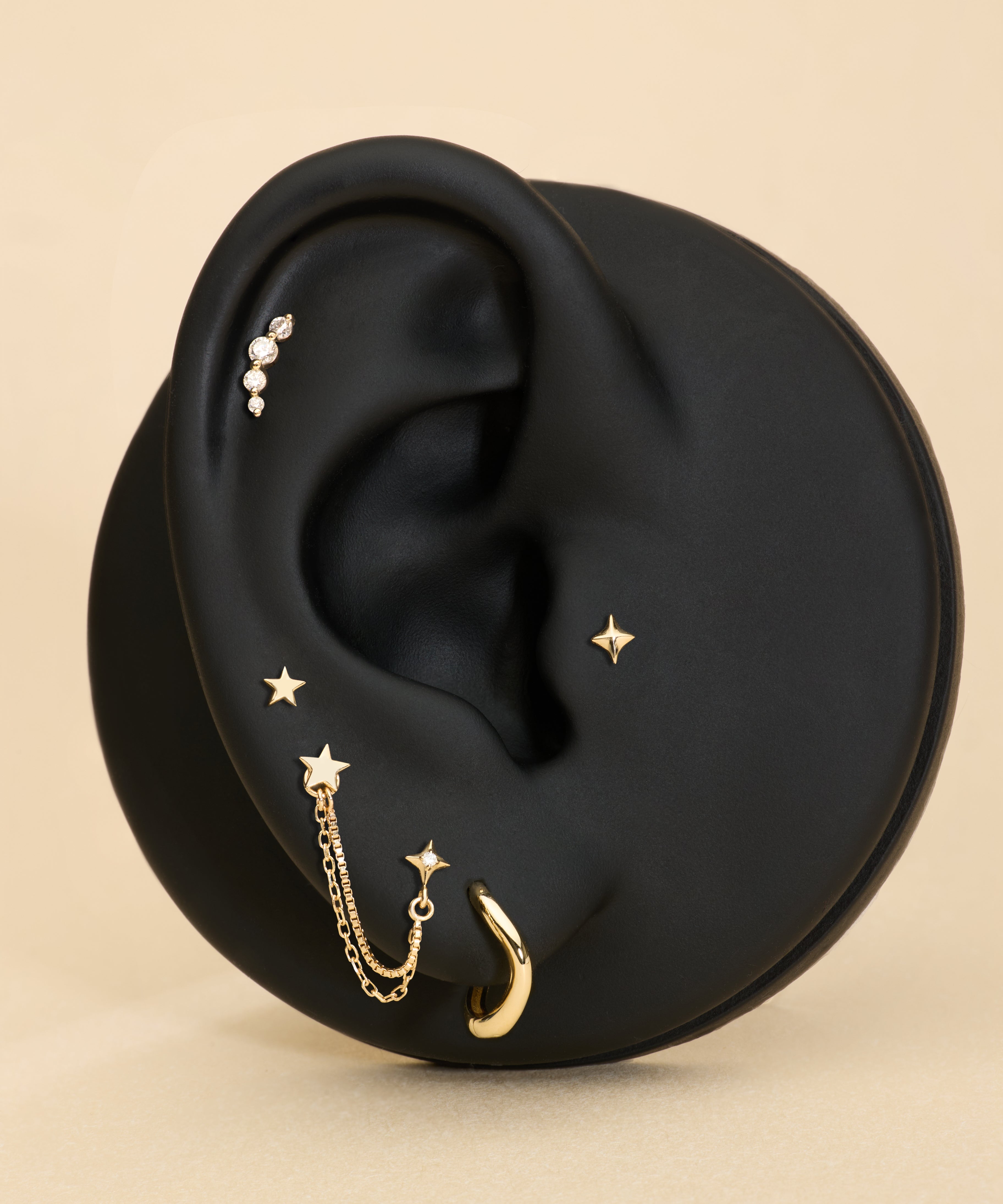 A black ear-shaped display showcases the 14K Solid Yellow Gold Four Point Star Flat Back Stud with other jewelry pieces against a beige background.
