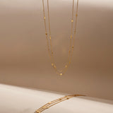 The Simona Double Chain Stacking Necklace in Gold features delicate double chains with small bead accents, displayed vertically and horizontally against a soft beige background.