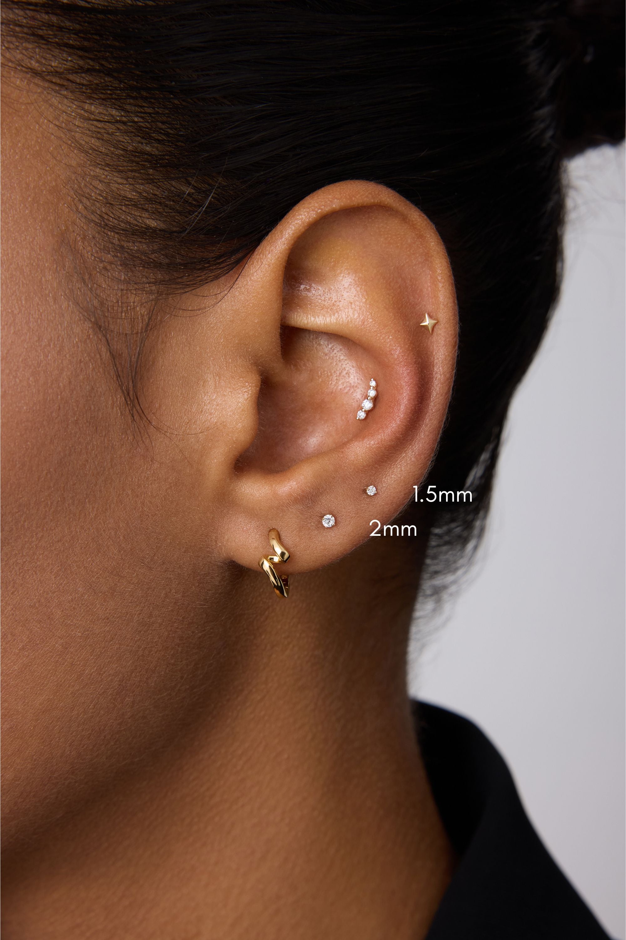 A close-up of a person’s ear with multiple piercings, featuring the 14K Solid Yellow Gold Lab-grown Diamond Prong Set Flat Back Stud - 1.5mm among other gold pieces. The person has medium brown skin and dark hair pulled back.