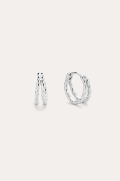 The Mae Twist Double Huggie Earrings in Silver feature a twisted, leafy design. Displayed on a plain background, one earring stands slightly open while the other lies flat, showcasing their intricate detailing.