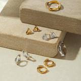 The Colette Droop Link Huggie Earrings in Gold are displayed on textured beige stone blocks, neatly arranged under soft lighting—perfect as stylish everyday earrings.