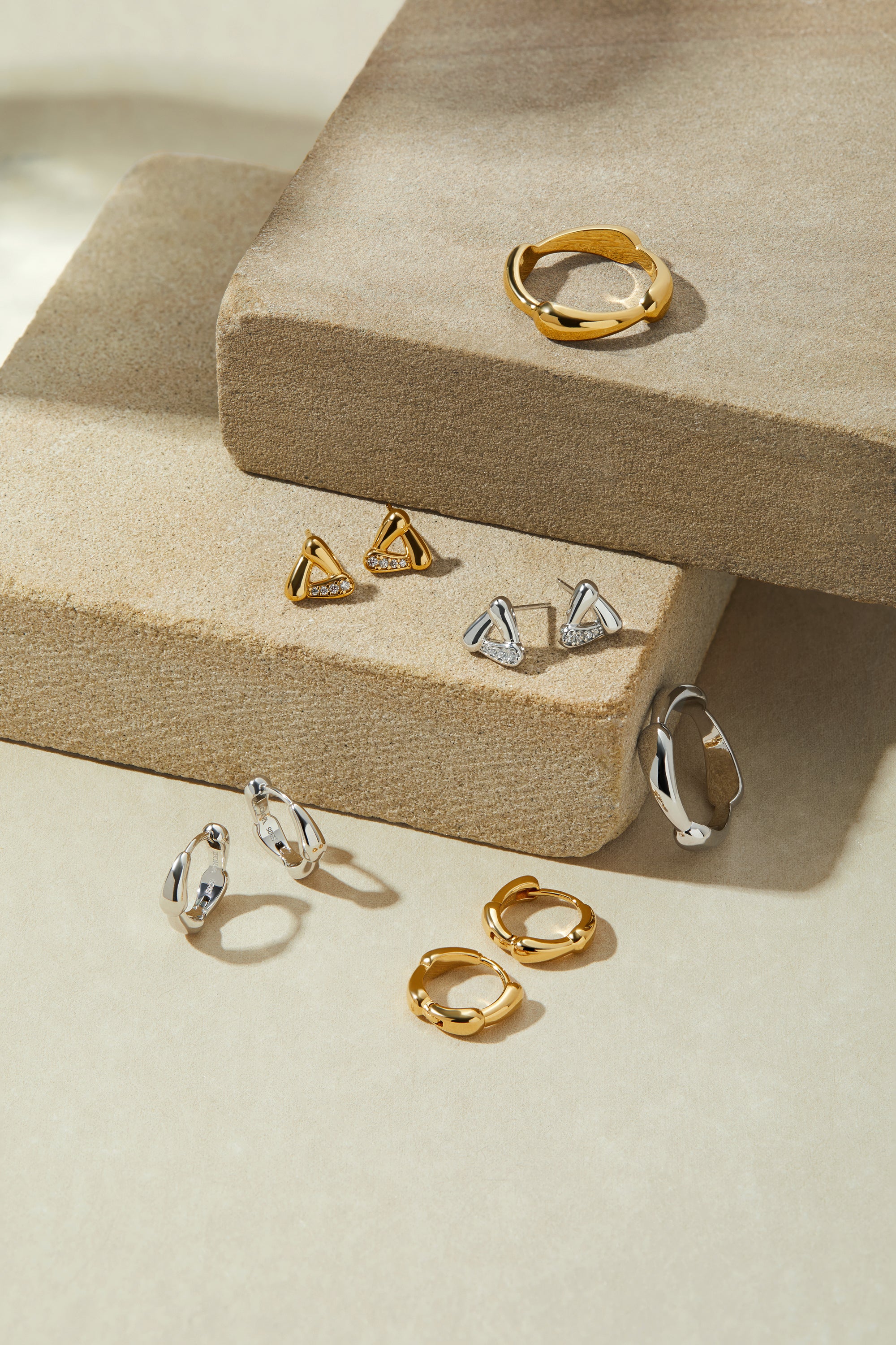 The Colette Droop Link Huggie Earrings in Gold are displayed on textured beige stone blocks, neatly arranged under soft lighting—perfect as stylish everyday earrings.